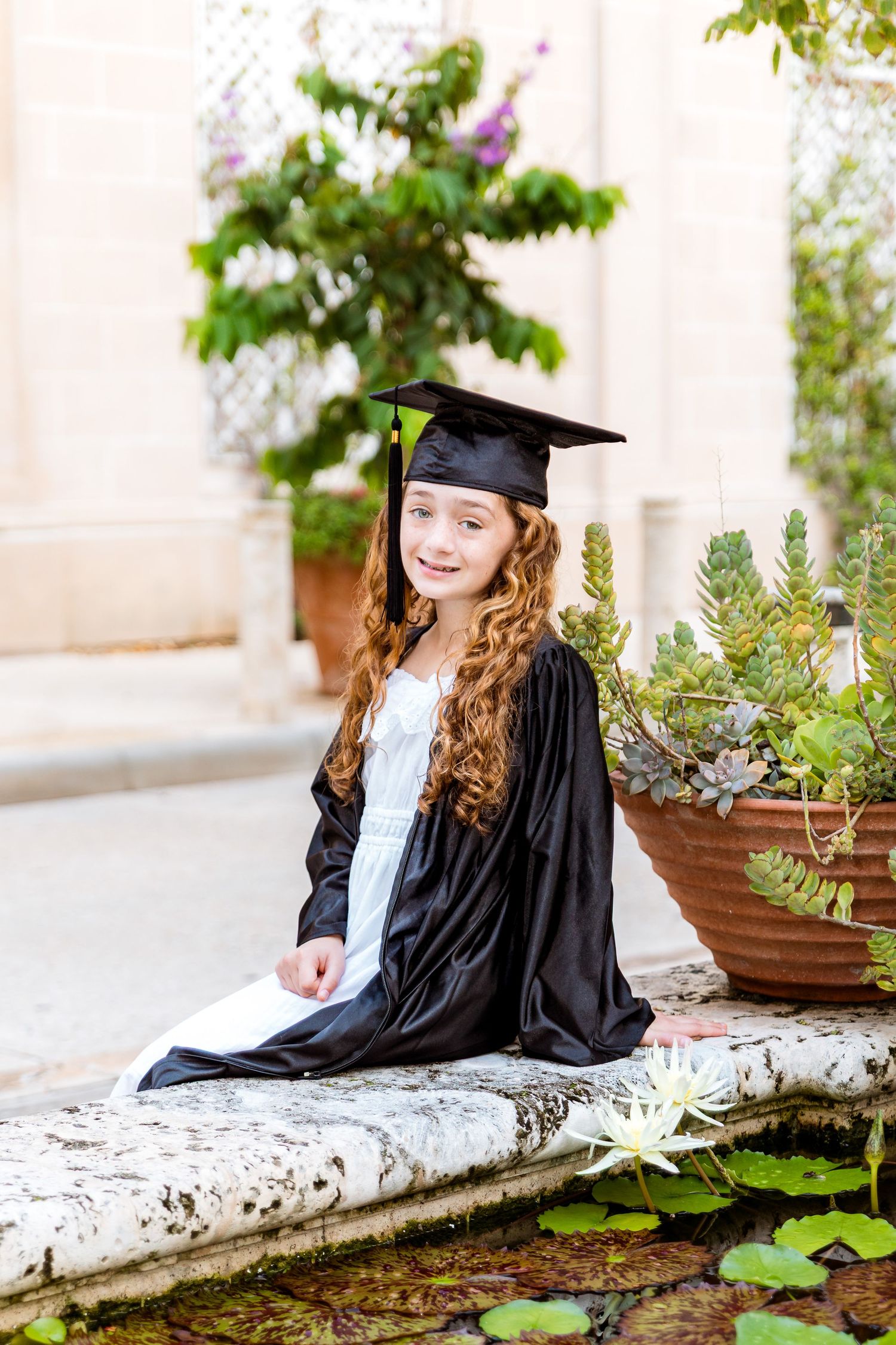 Young girl in black graduation cap and gown sits by garden fountain, smiling with long curly red hair.