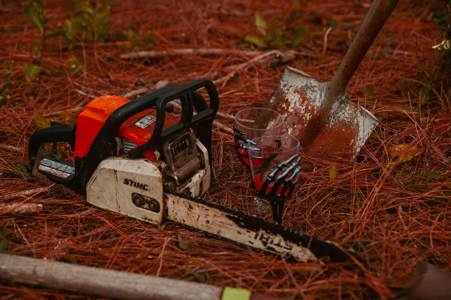 Multiple chainsaws and logging equipment lie scattered on red soil ground in a forest setting.