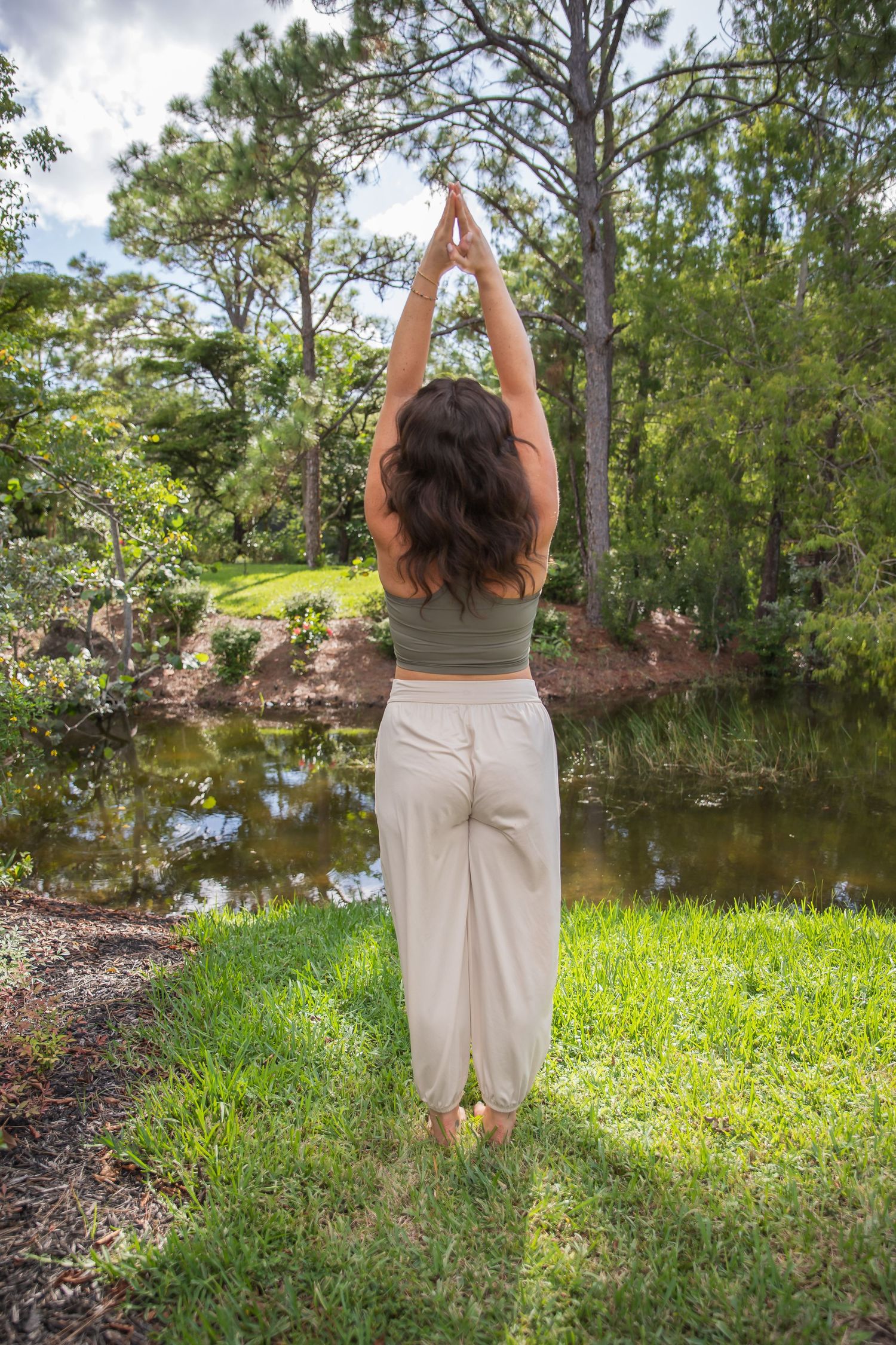 Someone practices yoga in a peaceful garden setting beside a pond surrounded by tall pine trees and lush grass.