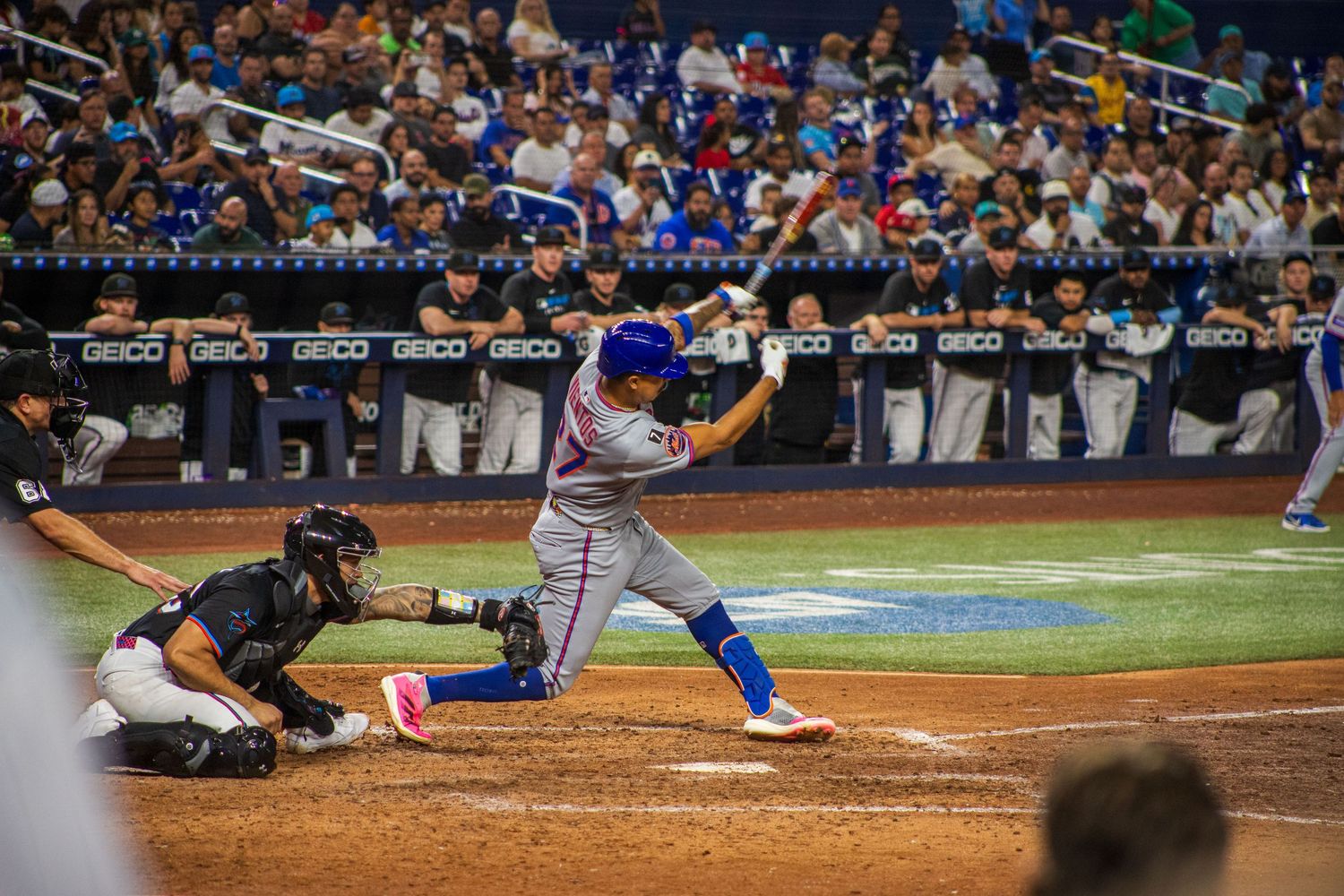 Baseball batter swings at pitch while catcher crouches behind home plate during night game.