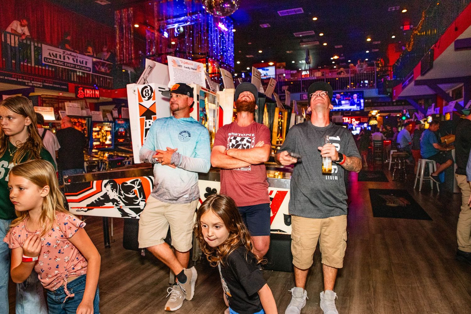 A lively scene of people, mostly young adults, gathered in a crowded arcade-style entertainment venue.