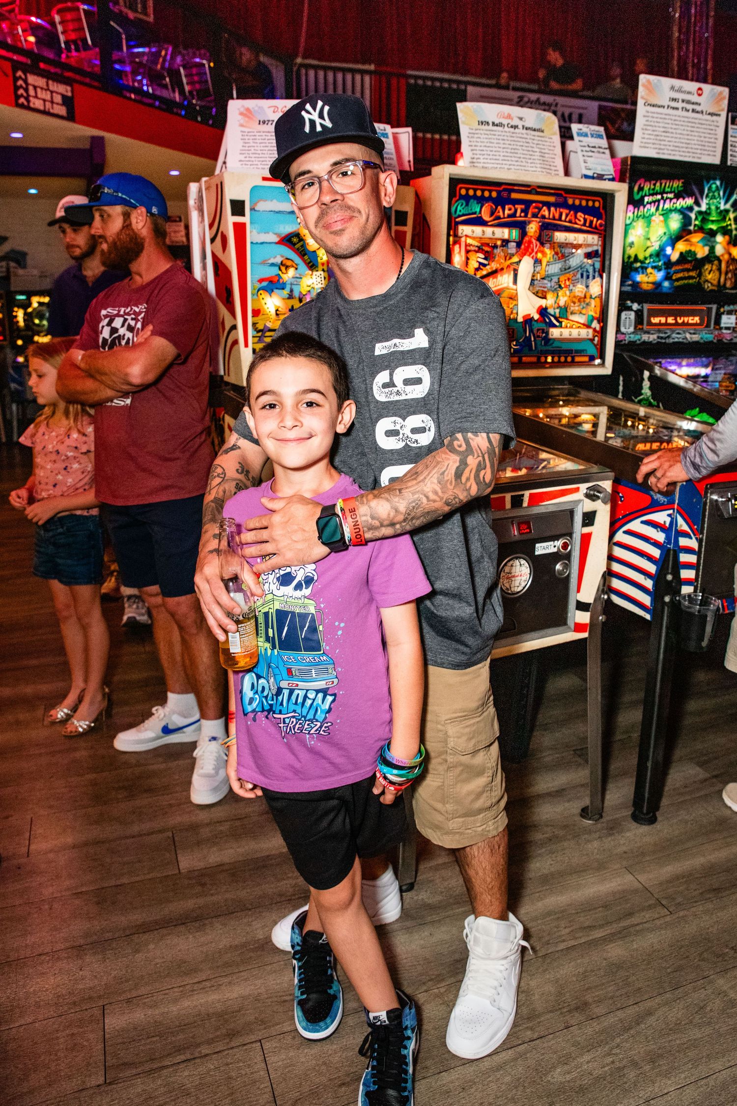 Tattooed man poses with young boy in front of vintage pinball machines at arcade.