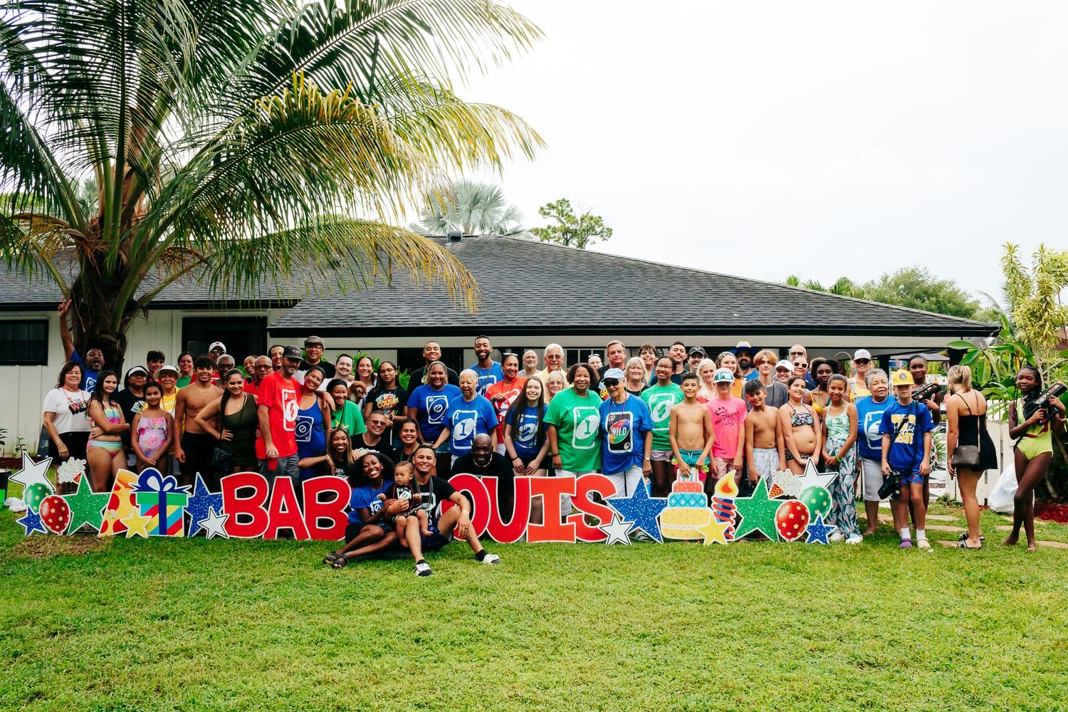 Large group photo of party guests gathered on a lawn with palm trees and colorful decorations.