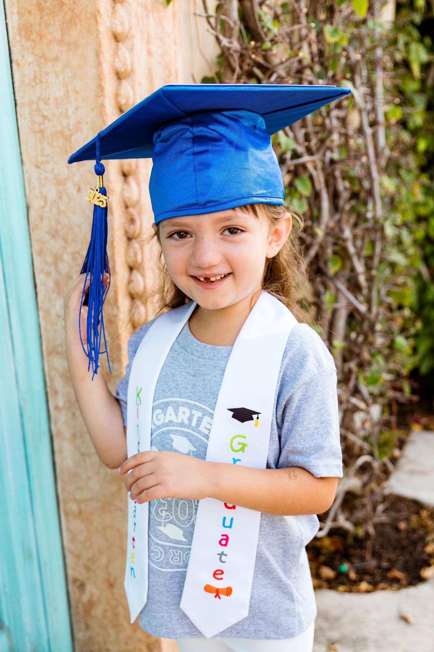 Young graduate in bright blue cap and decorated white sash stands outdoors in grey t-shirt by garden wall.