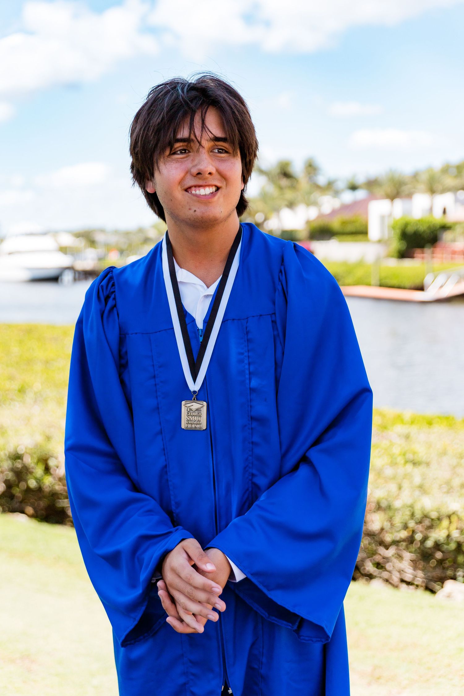 Graduate in bright blue cap and gown stands outdoors with medal around neck near marina on sunny day.
