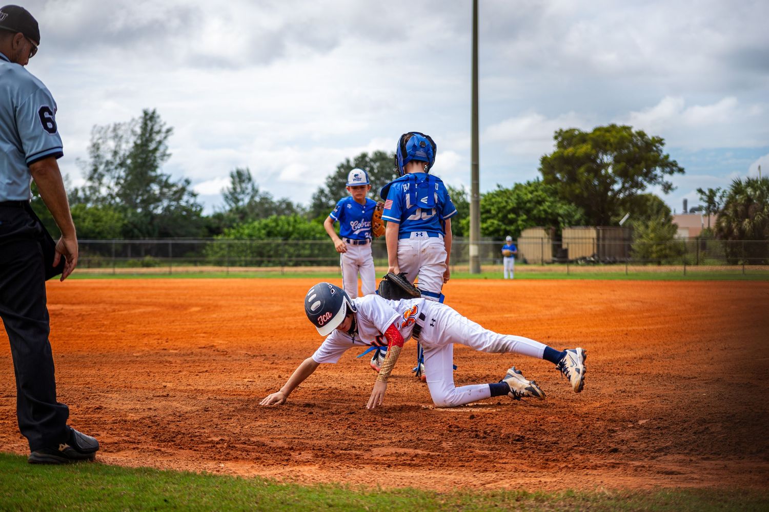 Baseball player slides into base creating a cloud of red dirt during a game on a cloudy day.