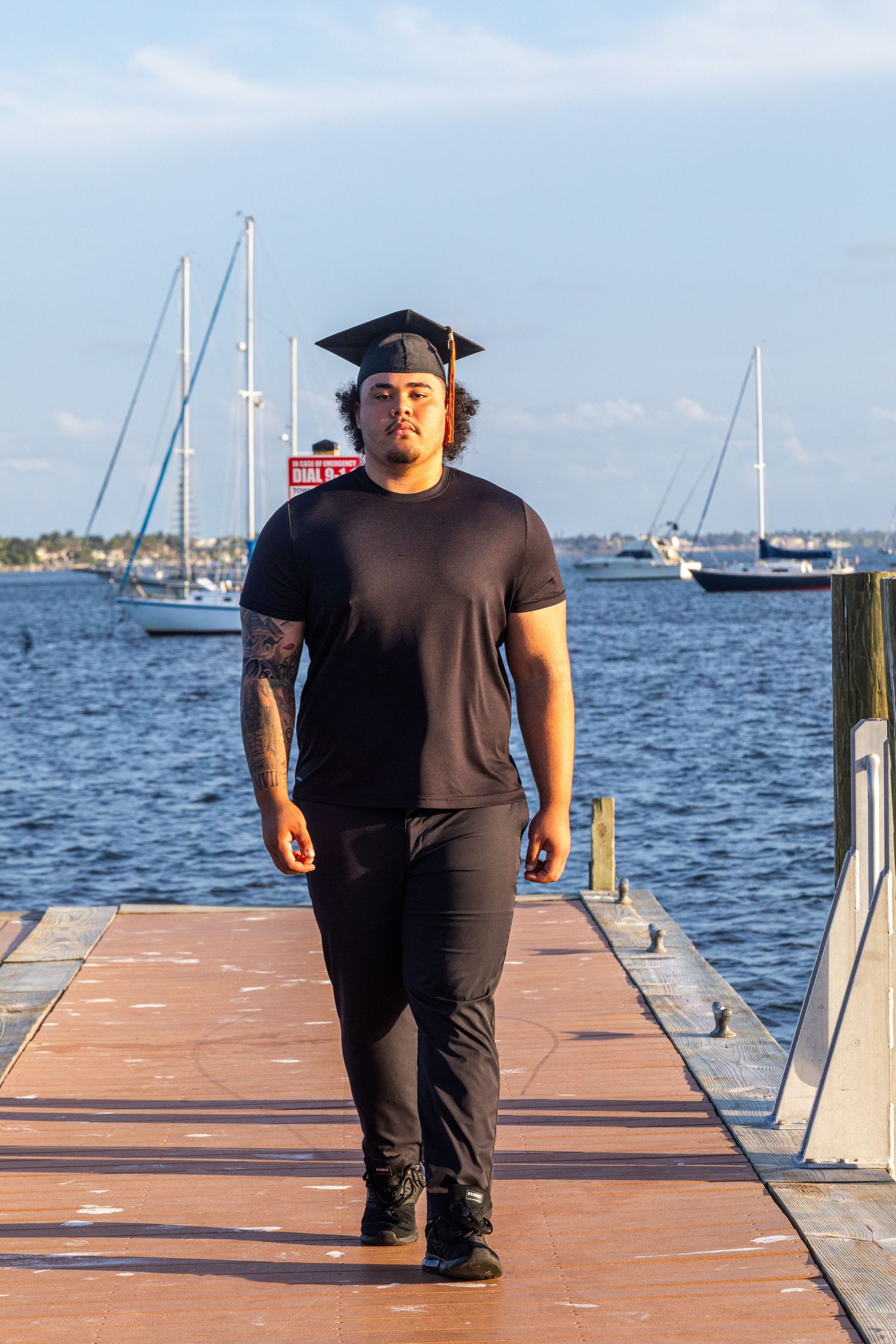 Young man in black outfit and graduation cap walks confidently on a waterfront dock with sailboats in the background.