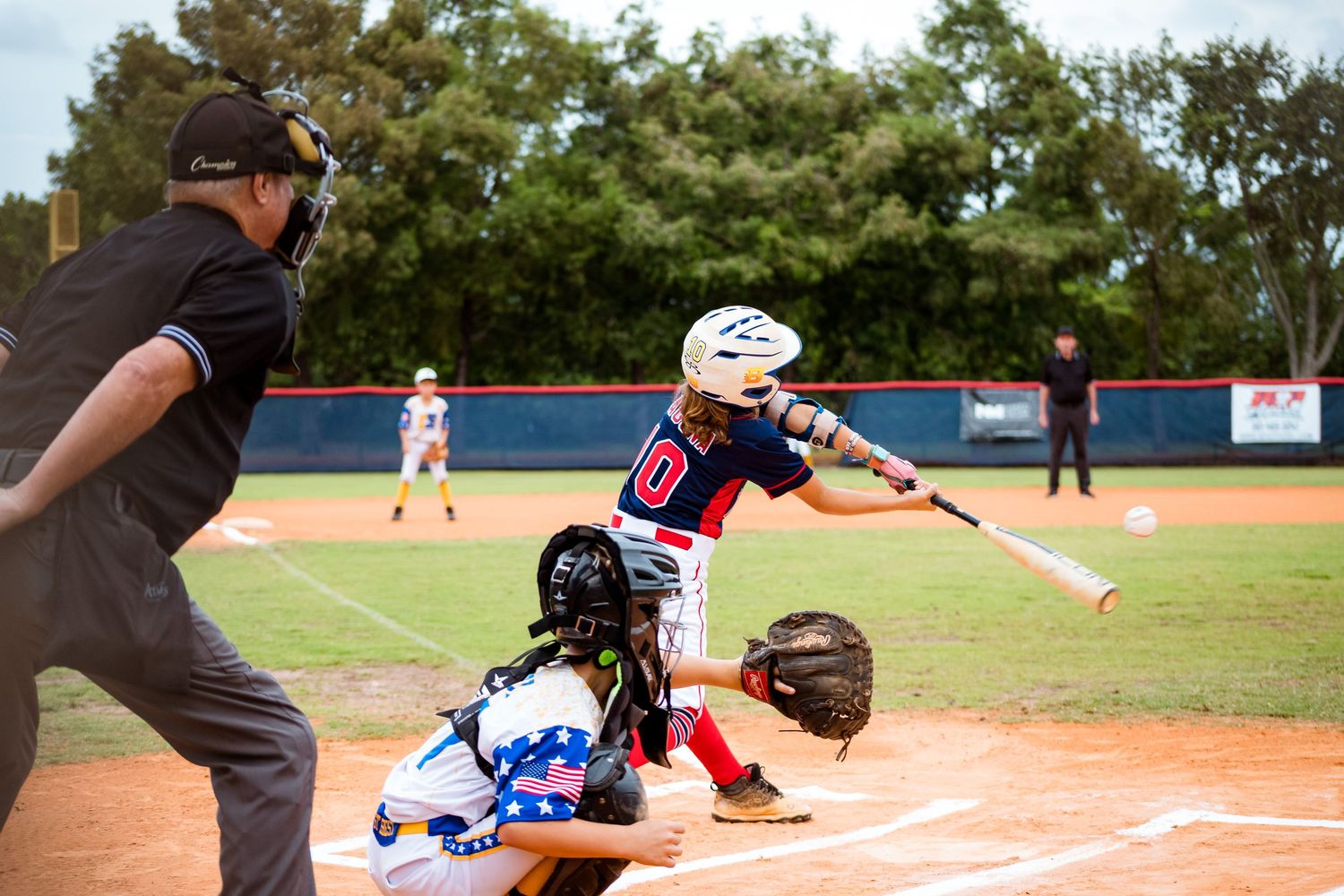 Baseball action shot at home plate with batter swinging and catcher crouching during a game on sunny day at ballpark.