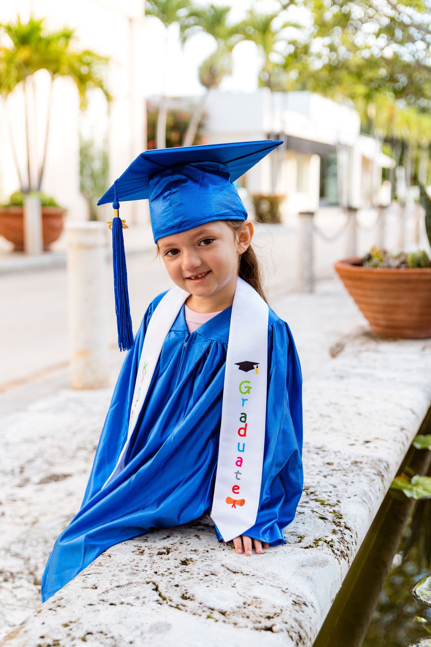 Graduate wearing bright blue cap and gown with decorated sash sits on stone ledge during outdoor graduation photoshoot.