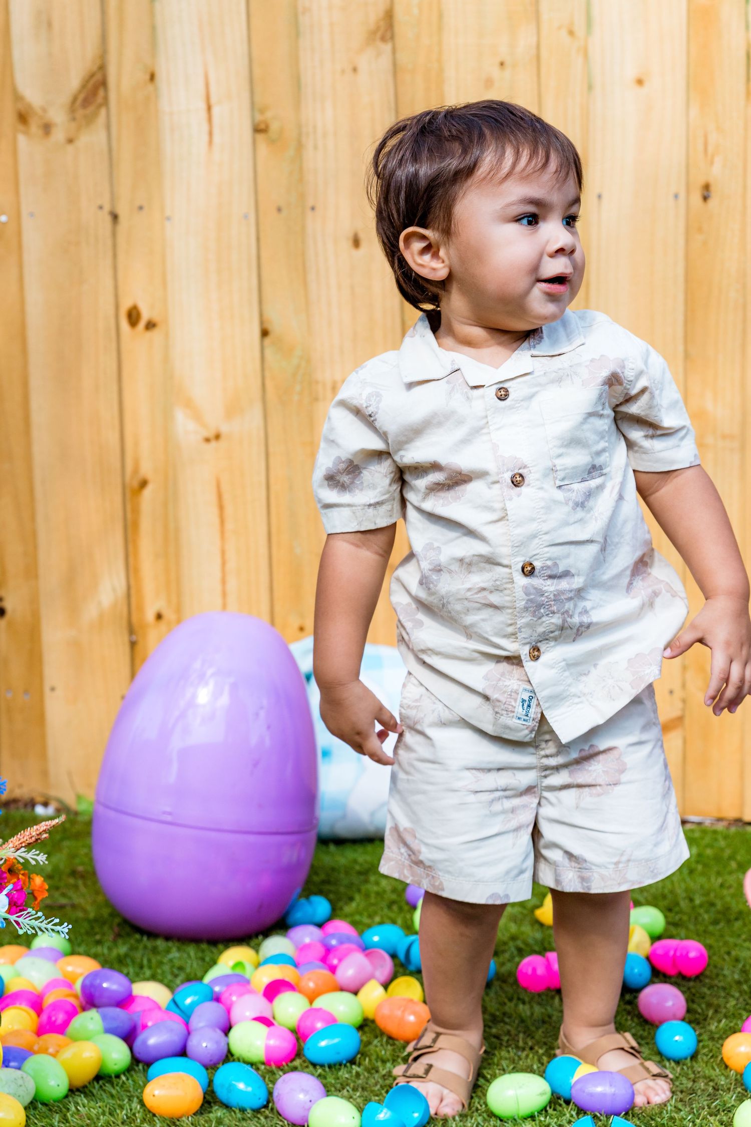 Small child in white outfit standing near large purple Easter egg and colorful plastic eggs.