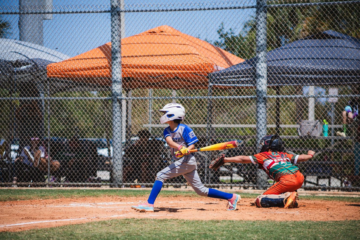 Action shot at baseball game showing batter in blue uniform swinging while catcher in red crouches at home plate.
