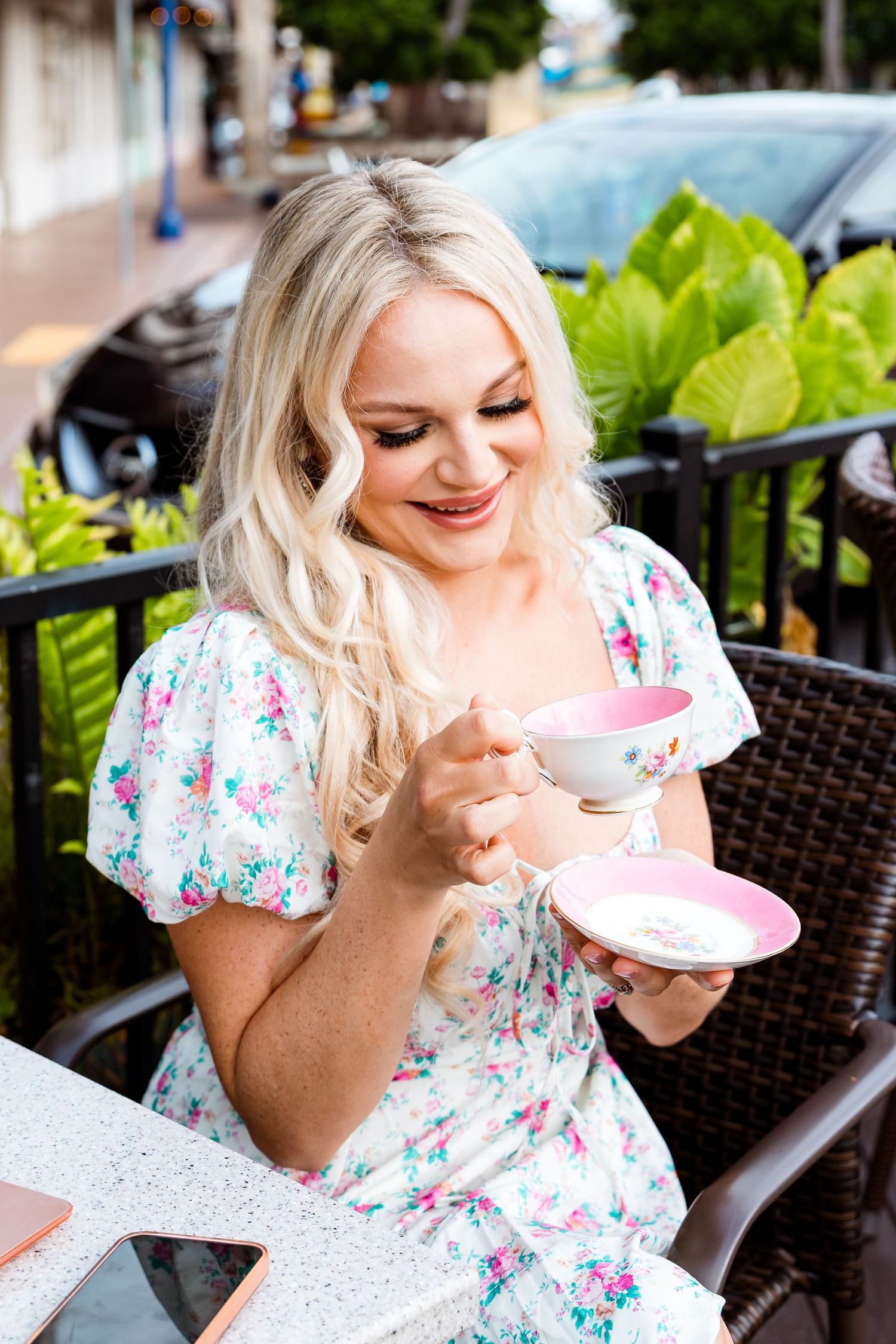 A person in a floral dress smiles while enjoying tea at an outdoor cafe patio.