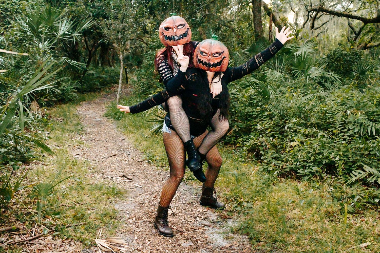 Two figures wearing pumpkin masks pose playfully on a forest path.