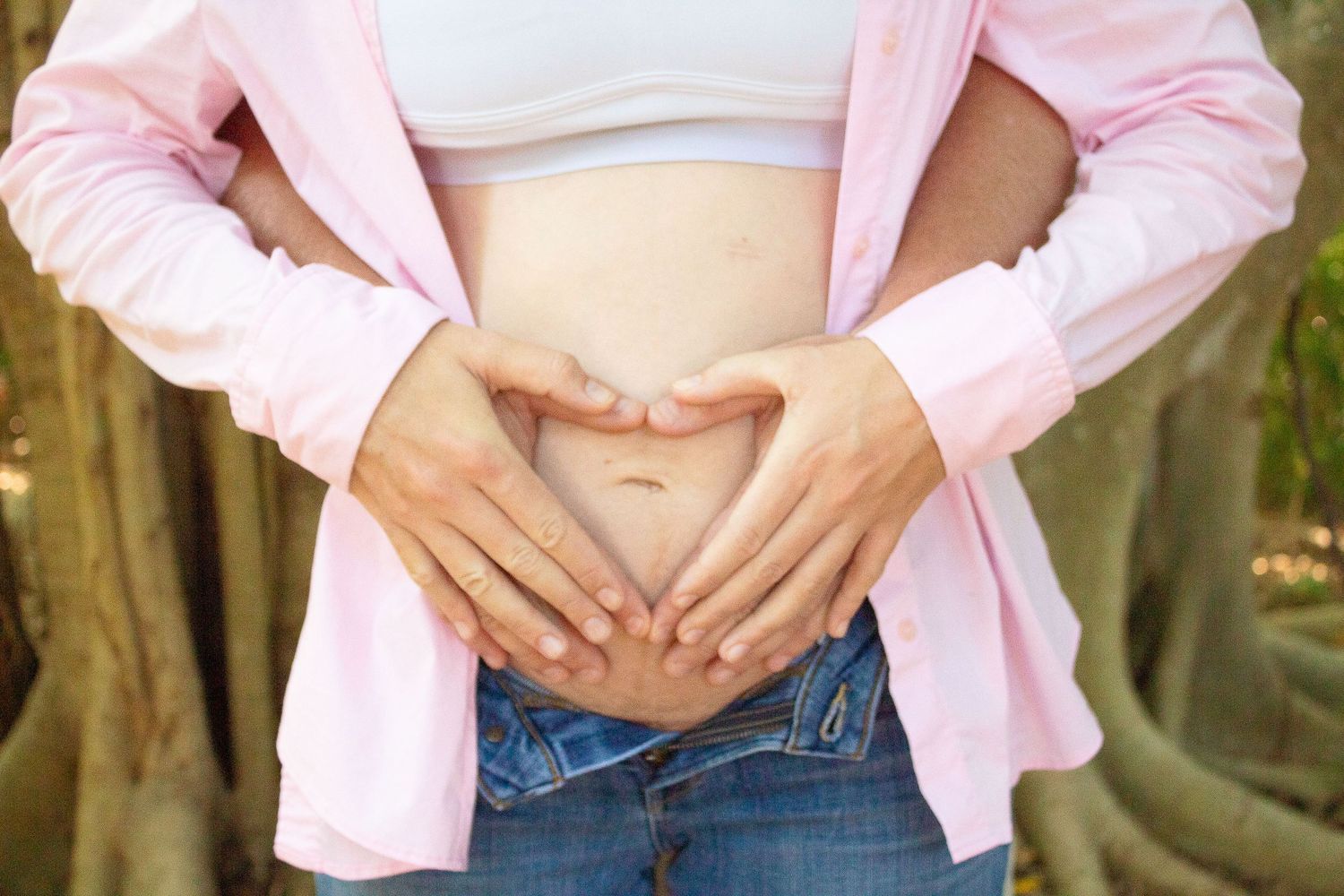 Close-up of hands forming heart shape over pregnant belly wearing pink cardigan and jeans.