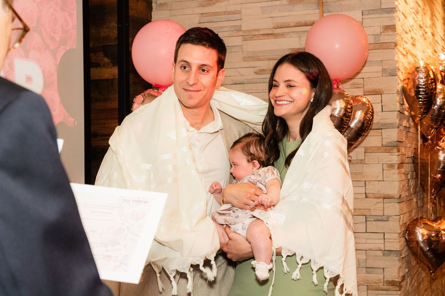 A ceremony moment with a couple wearing white and holding a baby, with pink balloons in the background.