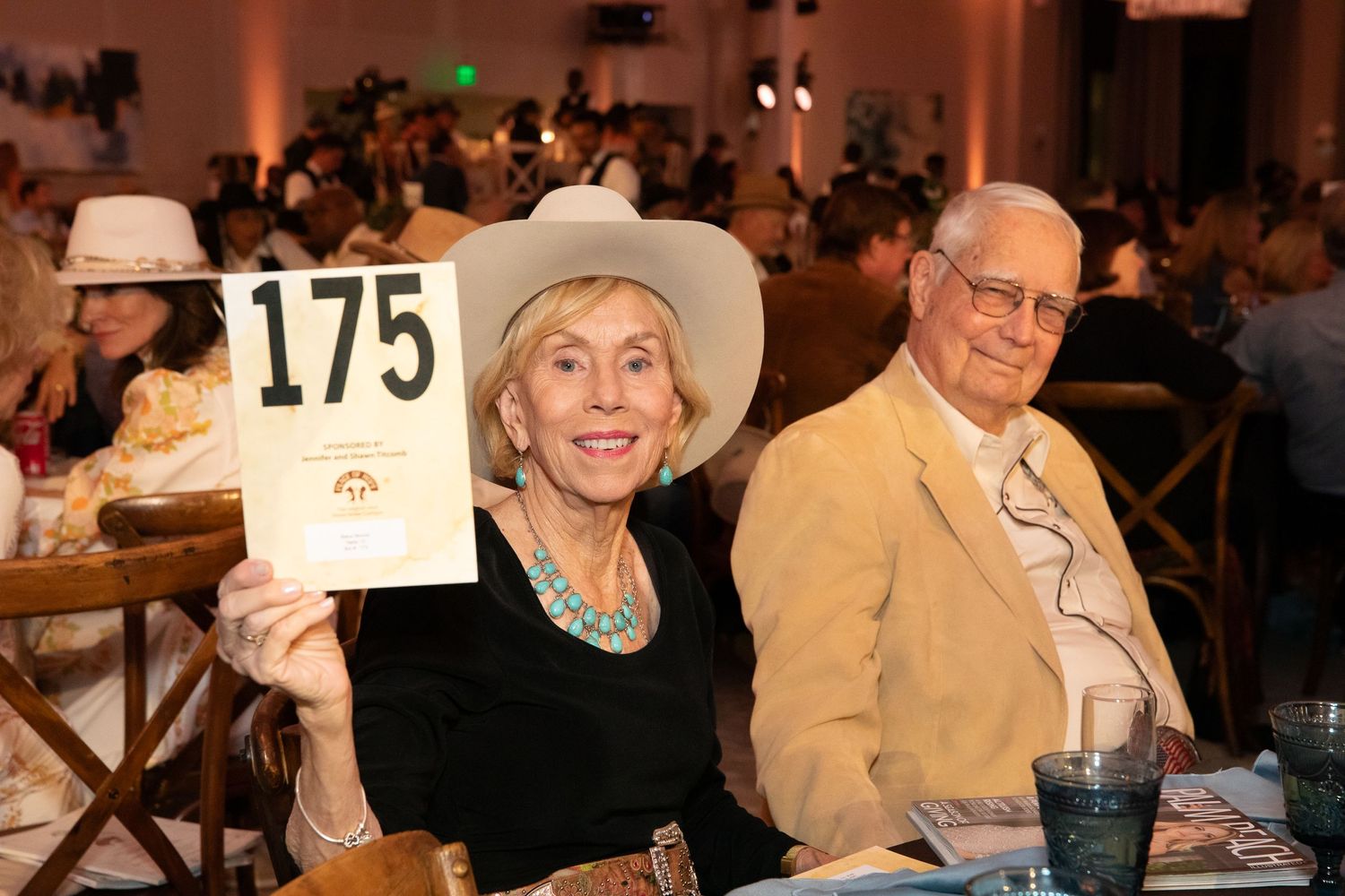Elderly couple in western attire holding auction paddle number 175 at formal event.