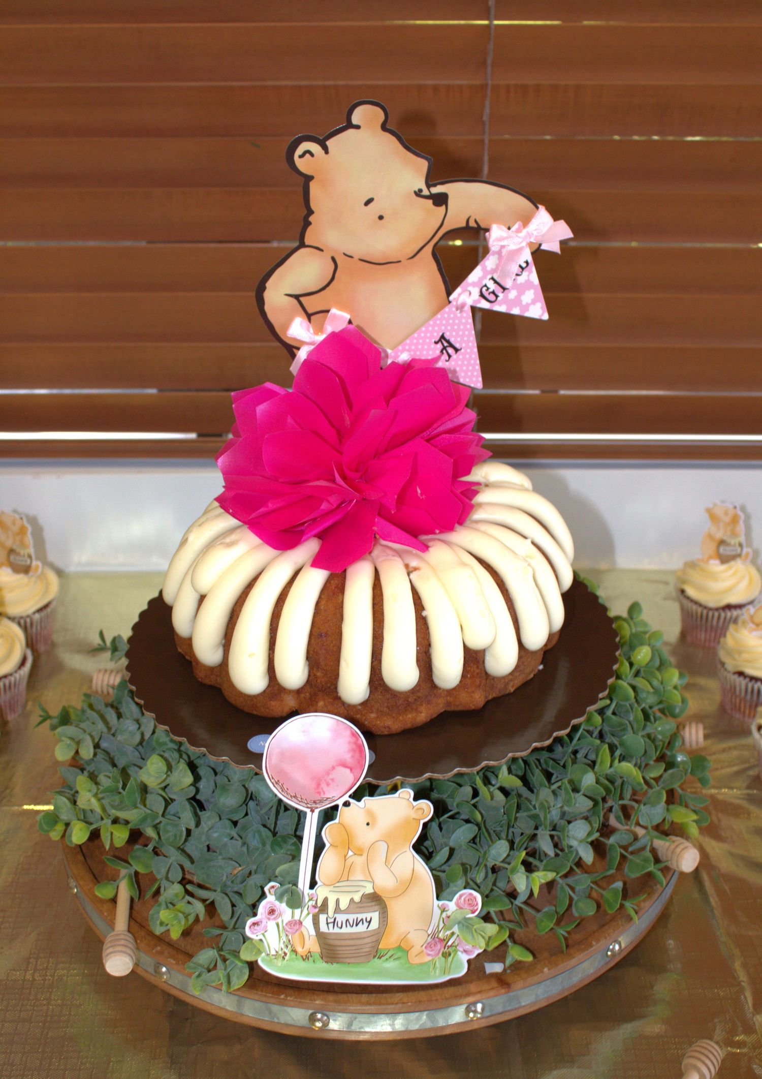 Bundt cake decorated with pink flower and Winnie the Pooh topper on decorative stand.