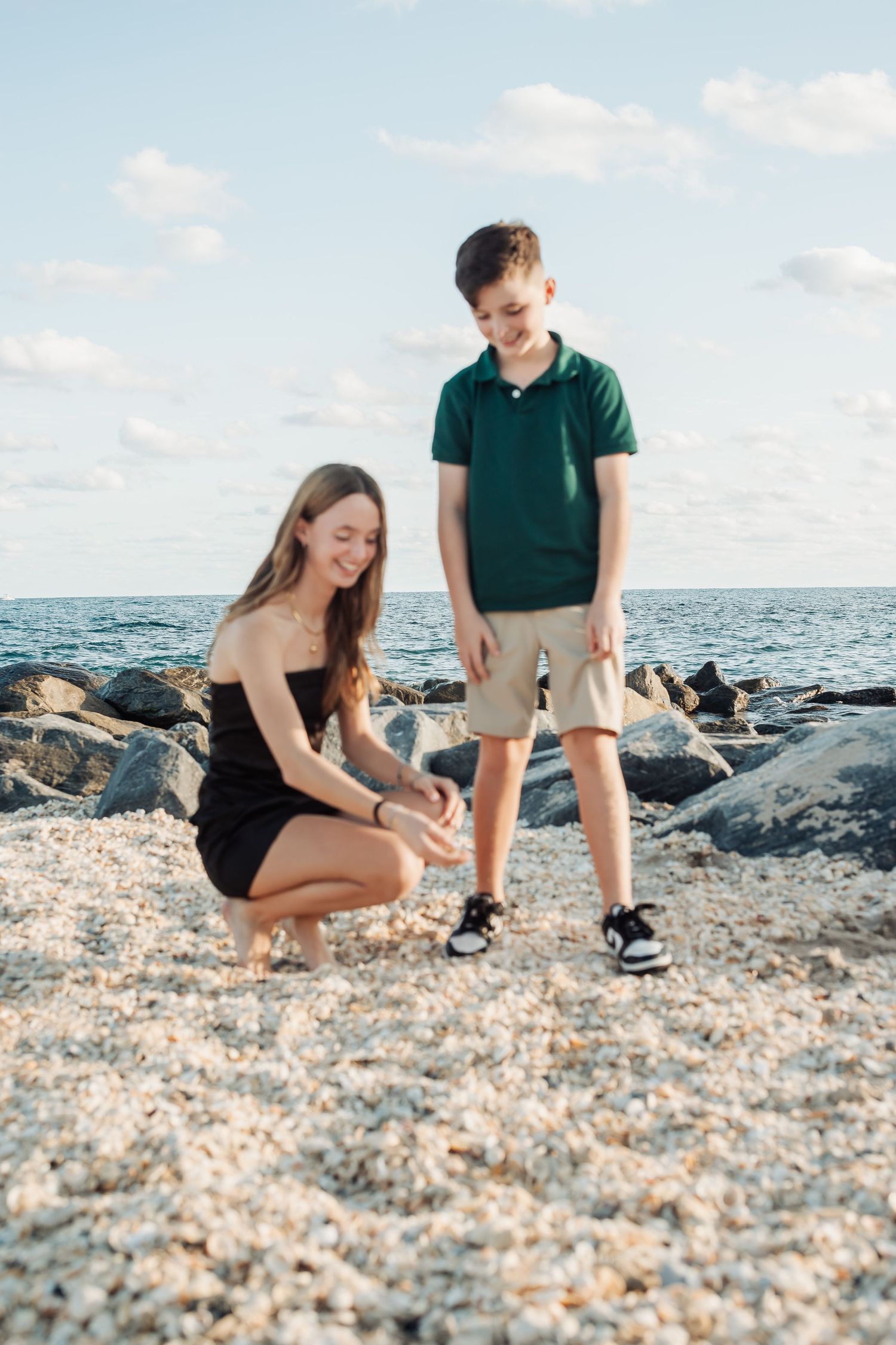 Two people exploring a rocky shoreline together against a bright blue ocean and sky backdrop.