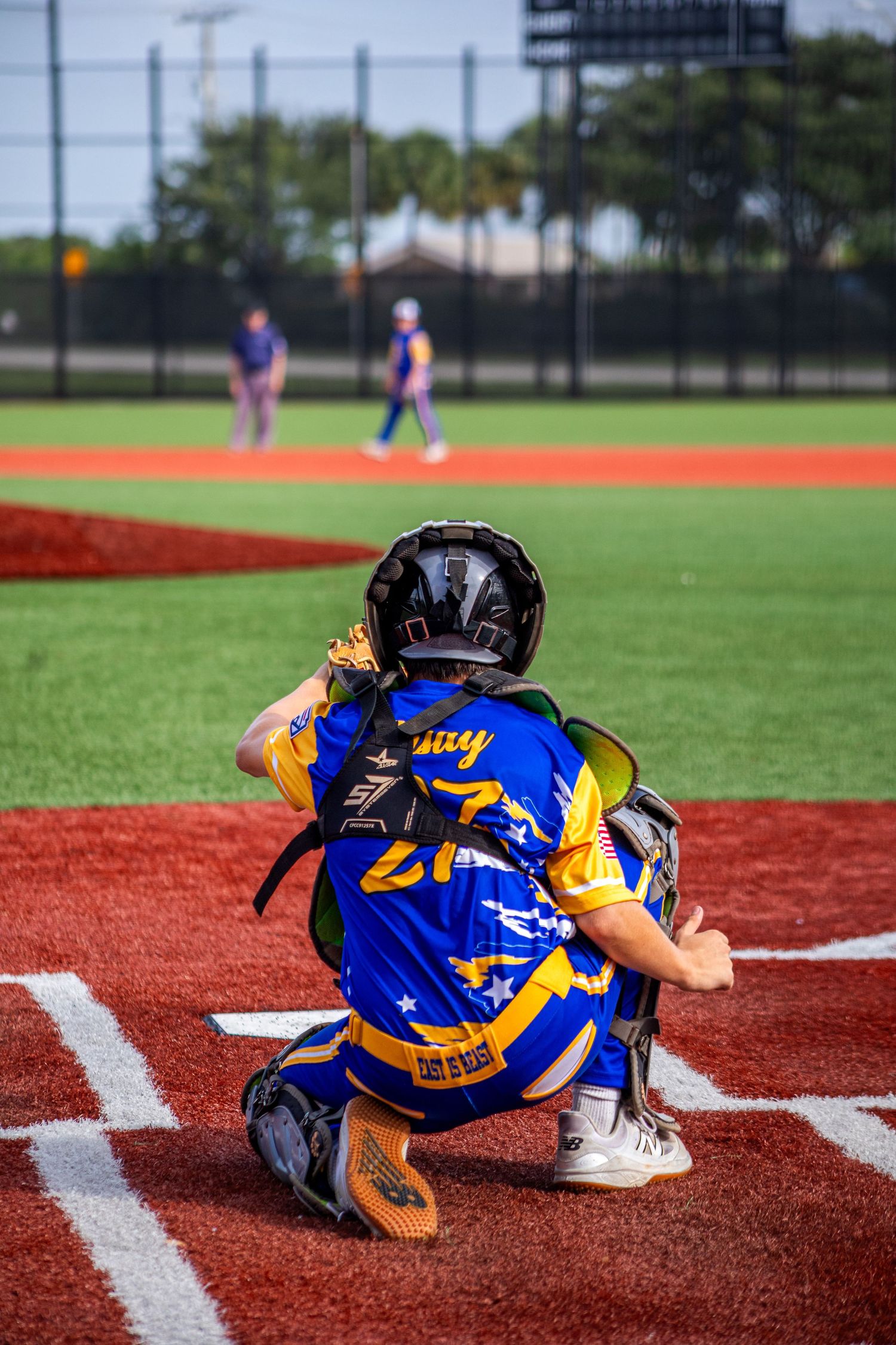 Youth baseball catcher in blue and gold uniform squatting behind home plate during a little league game.