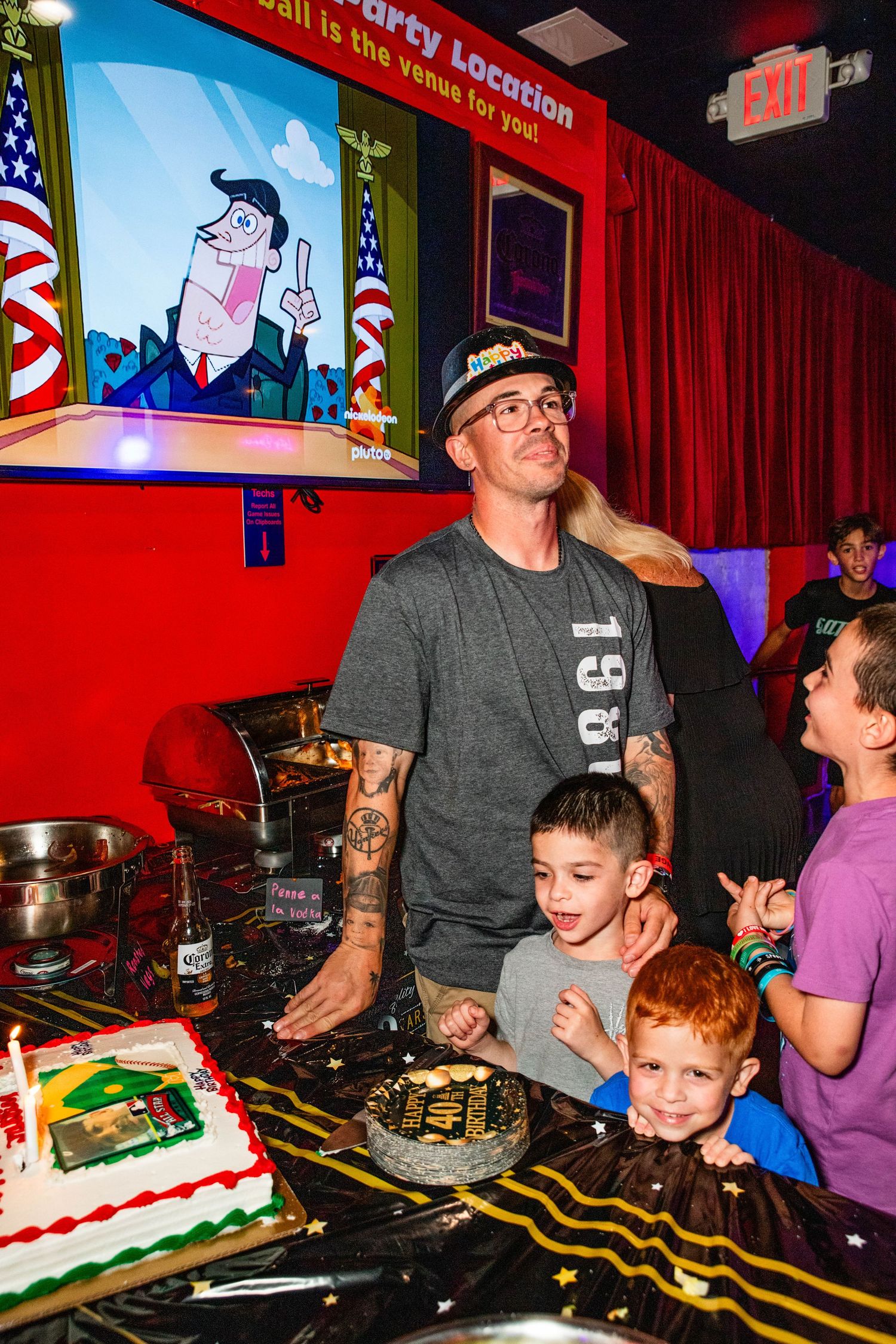 A group of people, including children, gathered around a table with a birthday cake and gaming equipment.