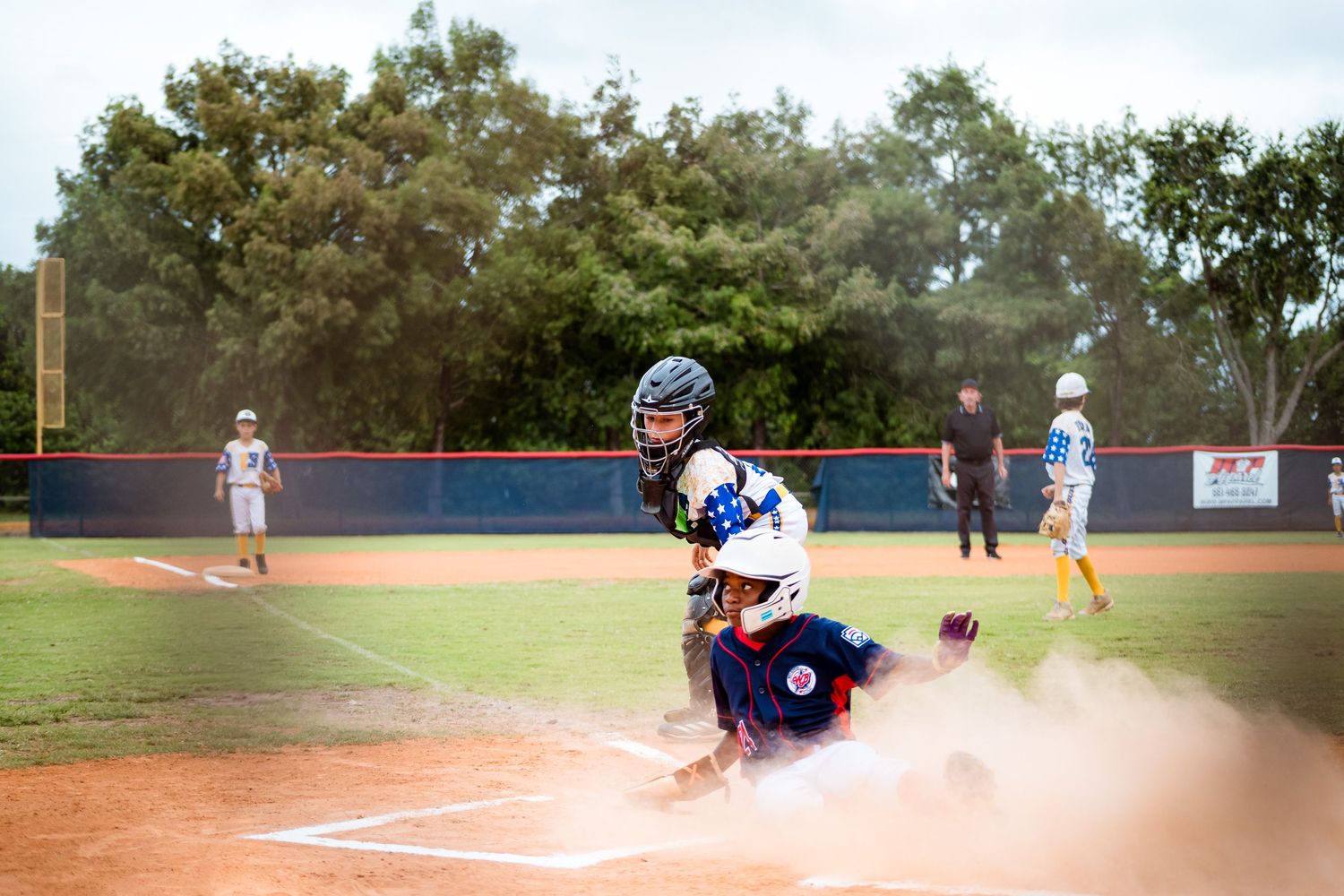 Baseball players in action during game as catcher crouches at home plate while dust kicks up from dirt.
