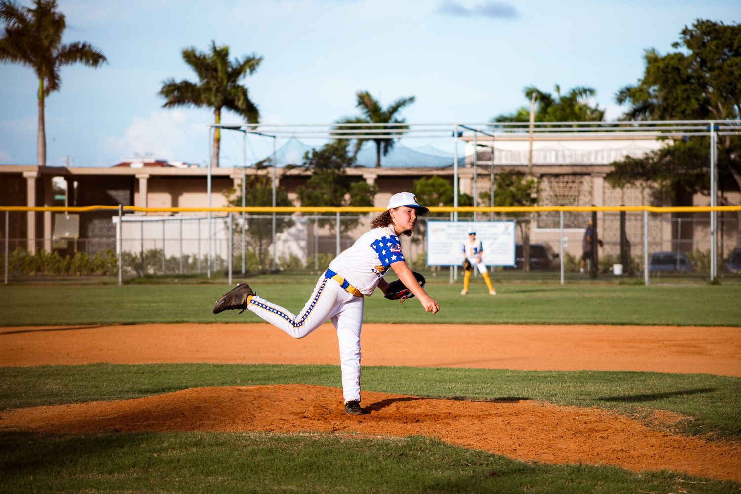Baseball player in blue uniform winds up for a pitch on a dirt mound with palm trees visible in the background.