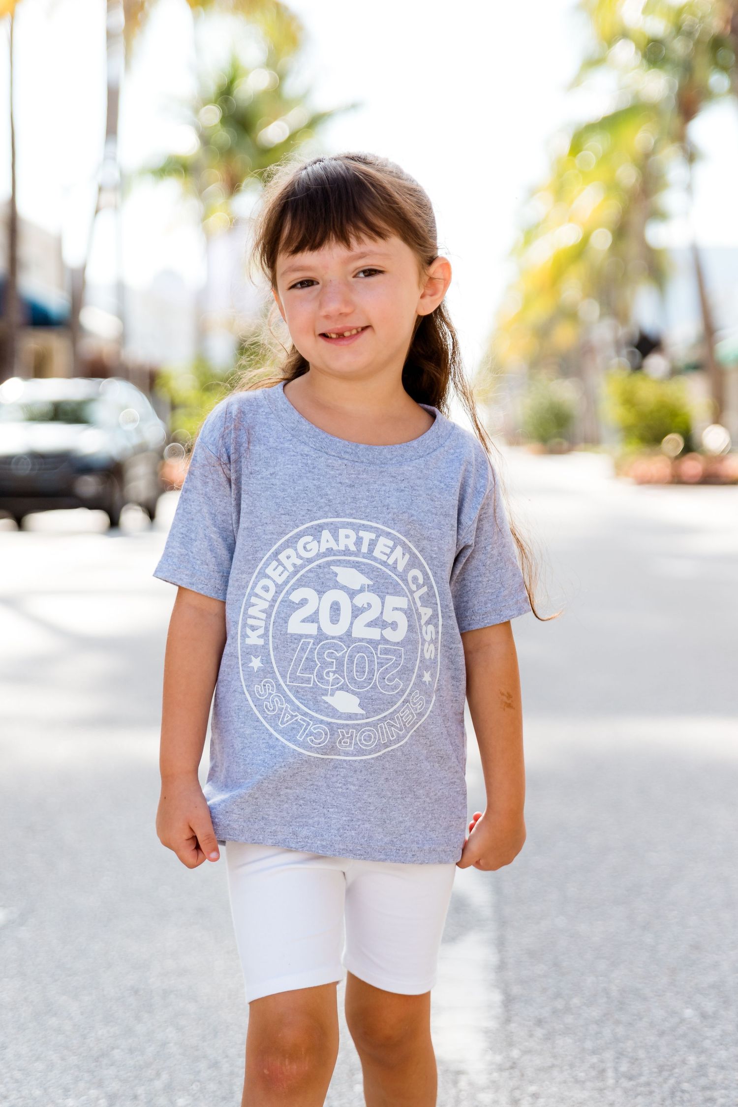 Young person wearing a gray graphic t-shirt and white shorts poses on a sunny palm-lined street.