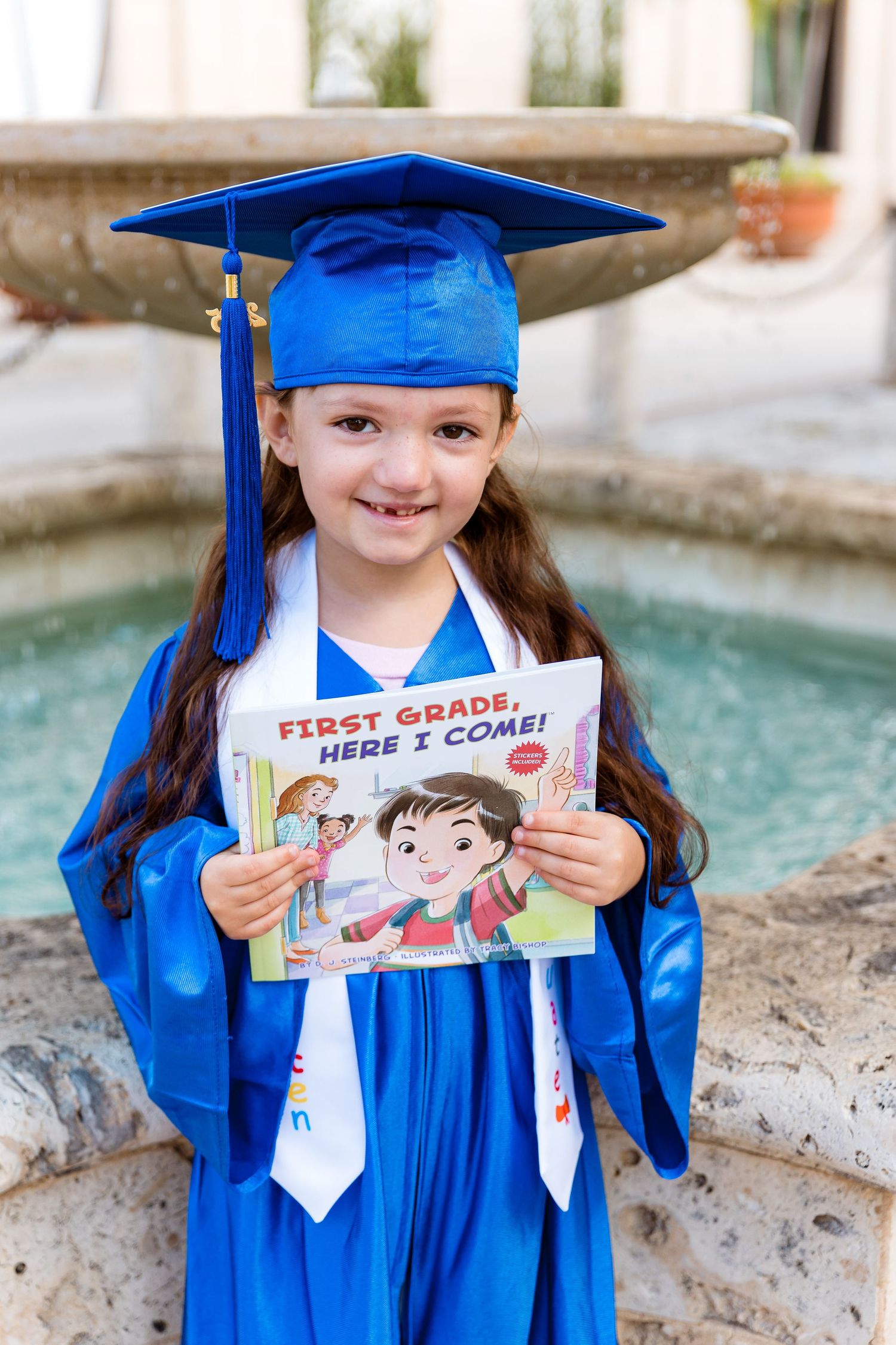 A young student wearing bright blue graduation regalia holds up a 'First Grade Here I Come' book by their fountain.