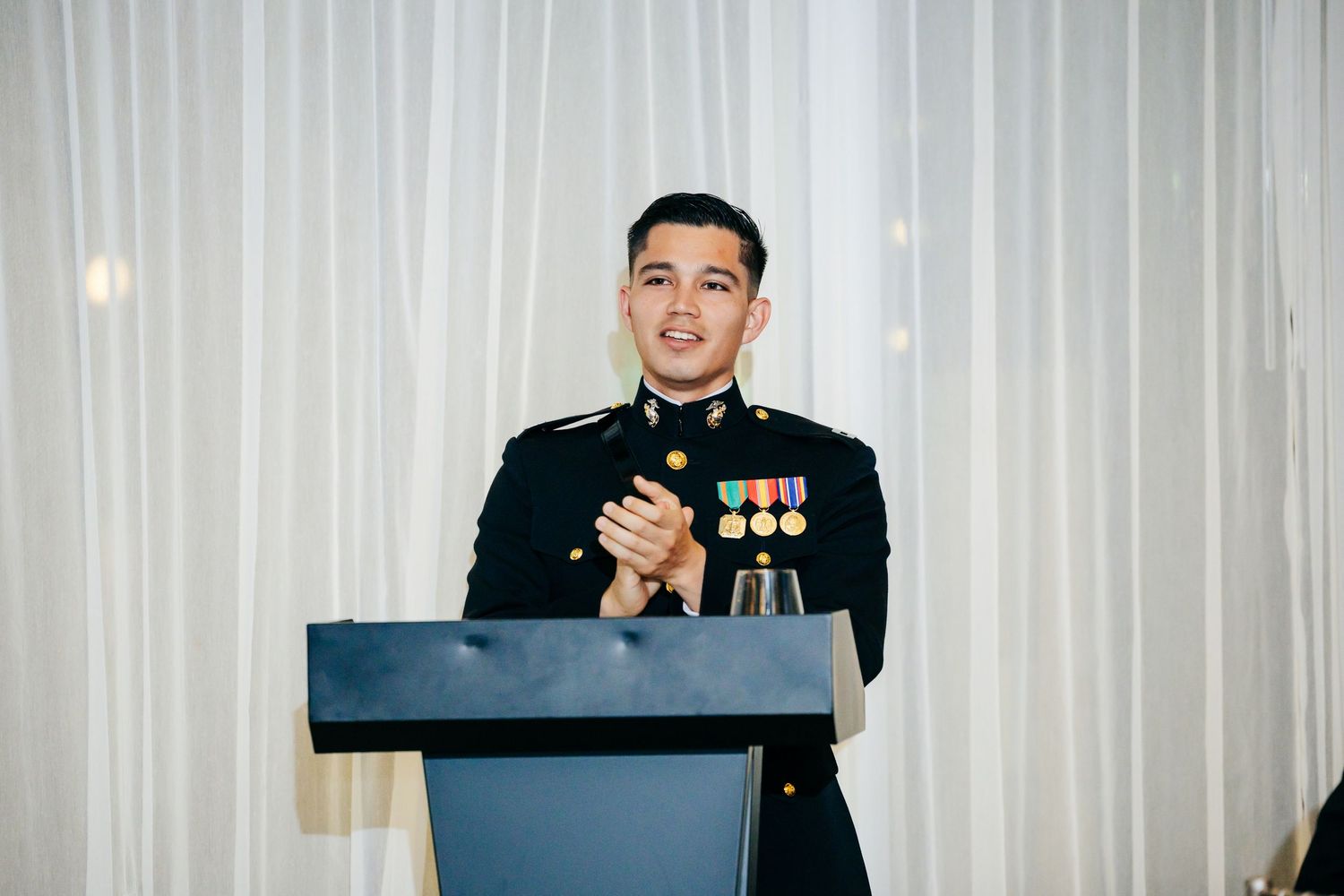 Military service members stand at a podium during a formal ceremony with white curtains in the background.