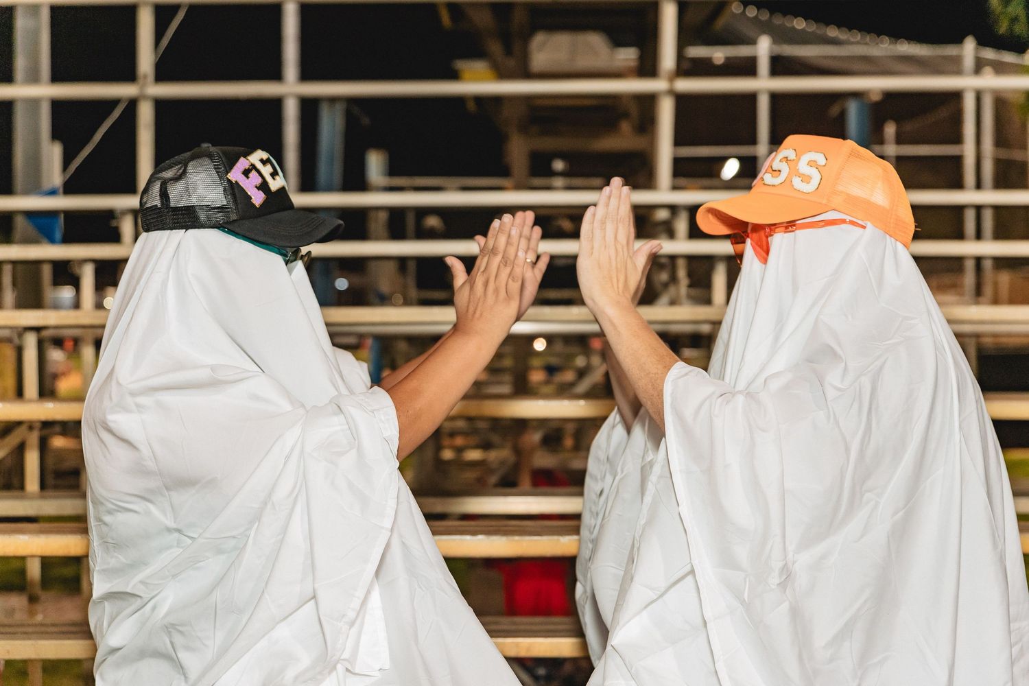 Group wearing white sheets raise their arms in unison while seated on outdoor bleachers.