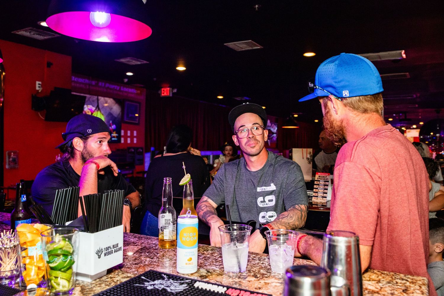 The image shows a group of people gathered around a bar, with one person wearing a blue construction helmet.