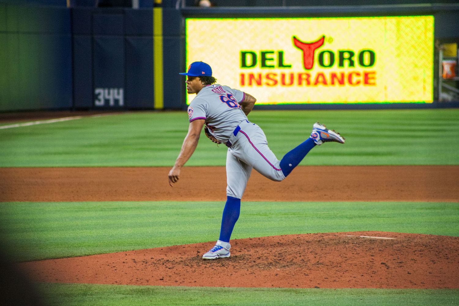 Pitcher follows through on delivery during night baseball game with illuminated Del Toro Insurance sign.
