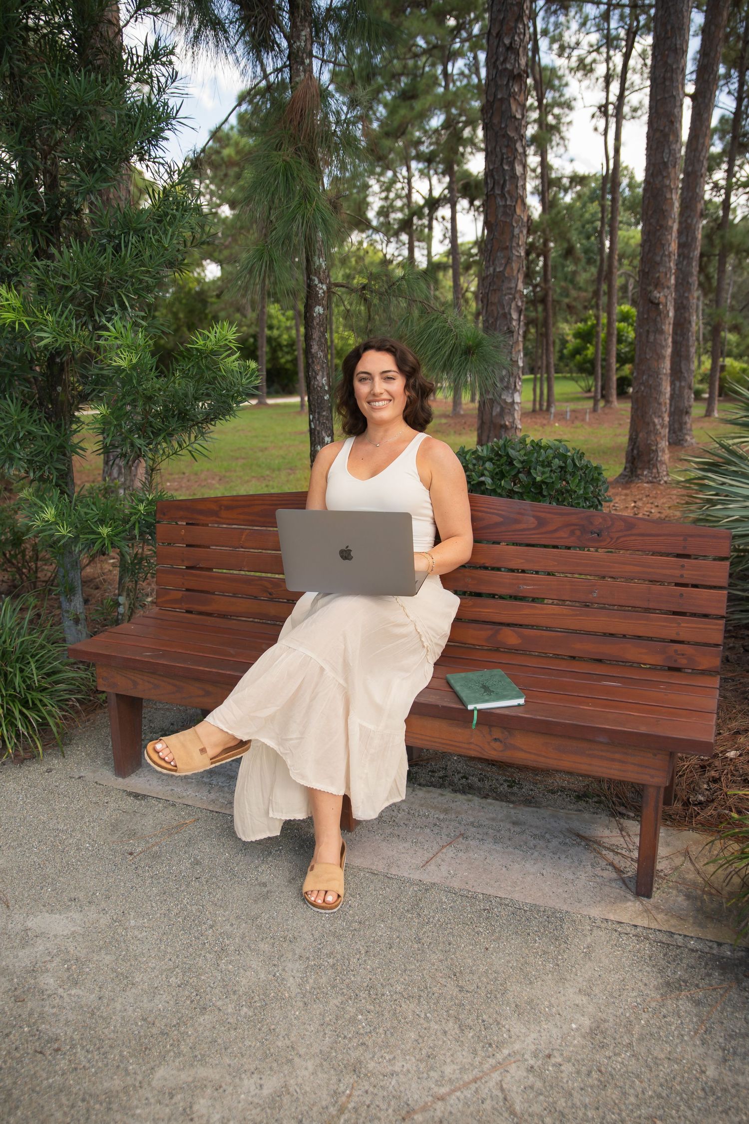 Person in white dress sitting and relaxing on wooden bench surrounded by pine trees.