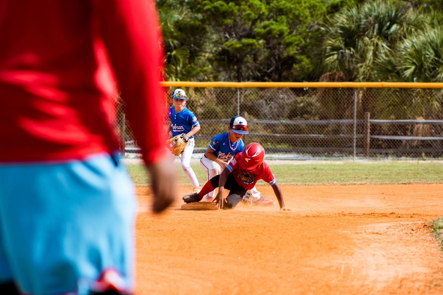 Youth baseball game in action on a dusty infield with players in red and blue uniforms.