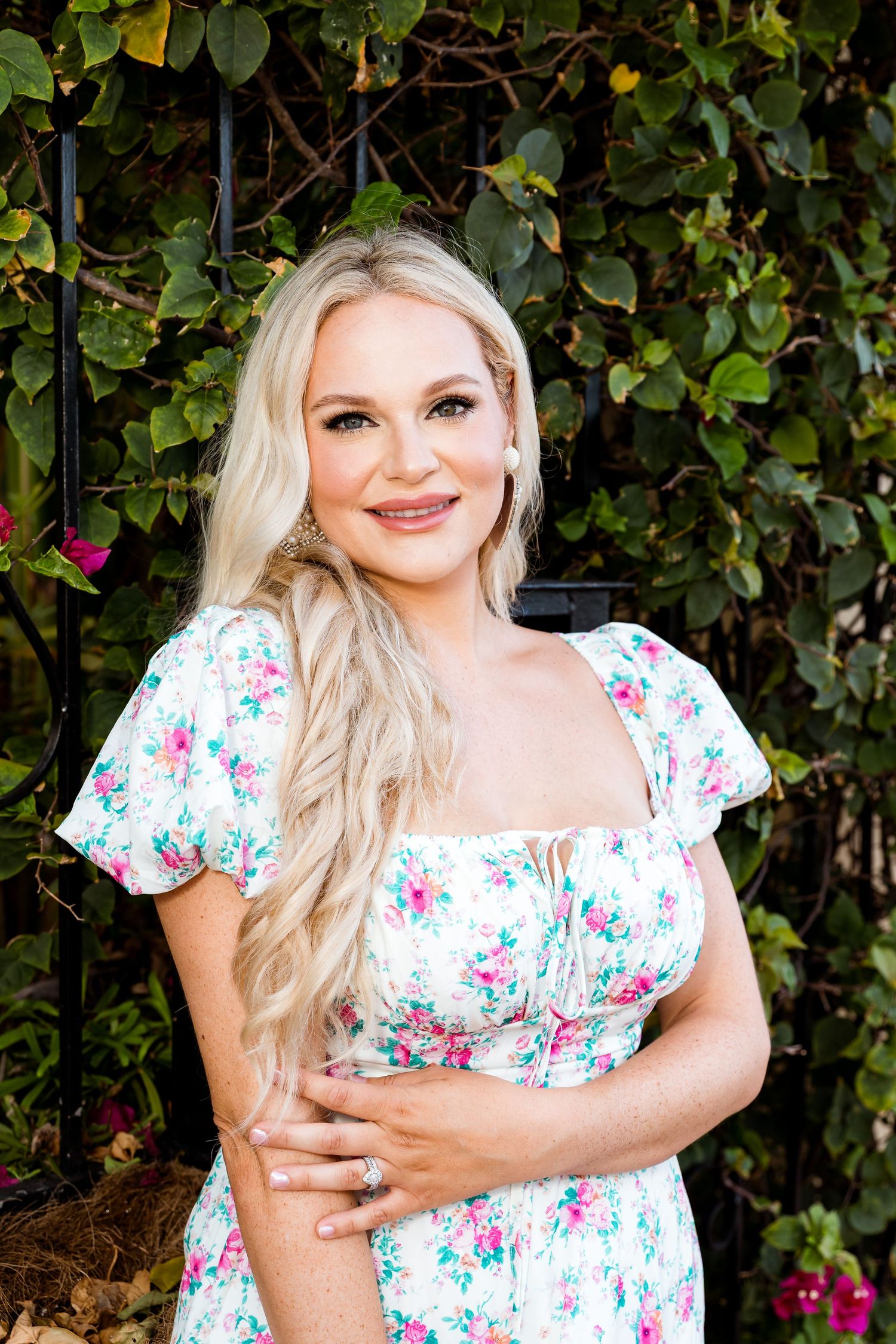Woman in white floral dress posing against vine-covered wall with pink flowers.