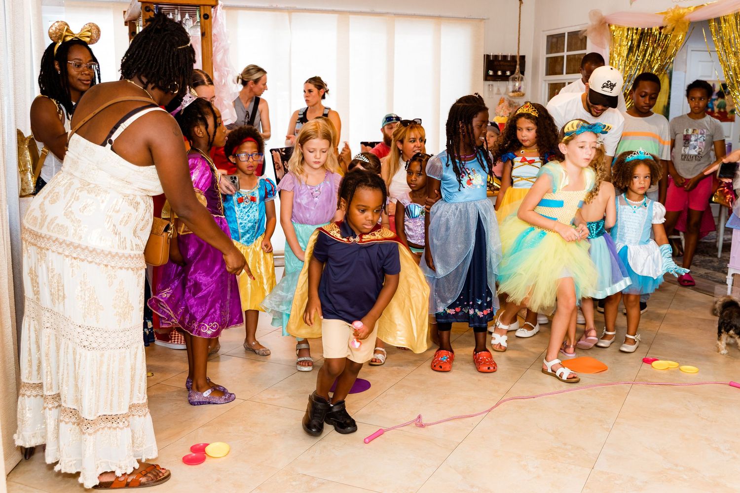 A group of children in colorful party dresses dance together during a festive celebration with rose petals on the floor.
