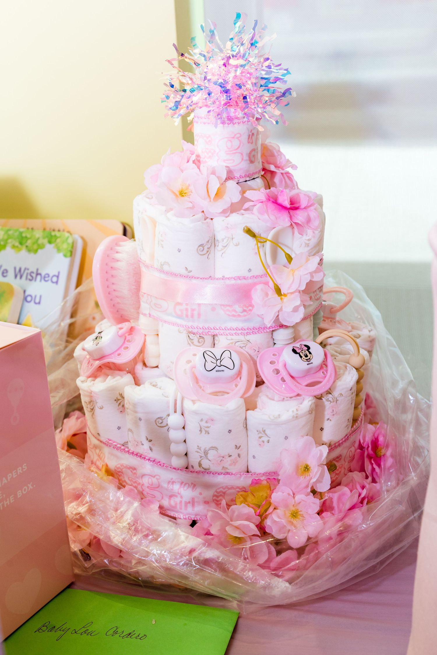 Pink themed baby shower cake decorated with flowers and delicate ornaments on a white tablecloth.