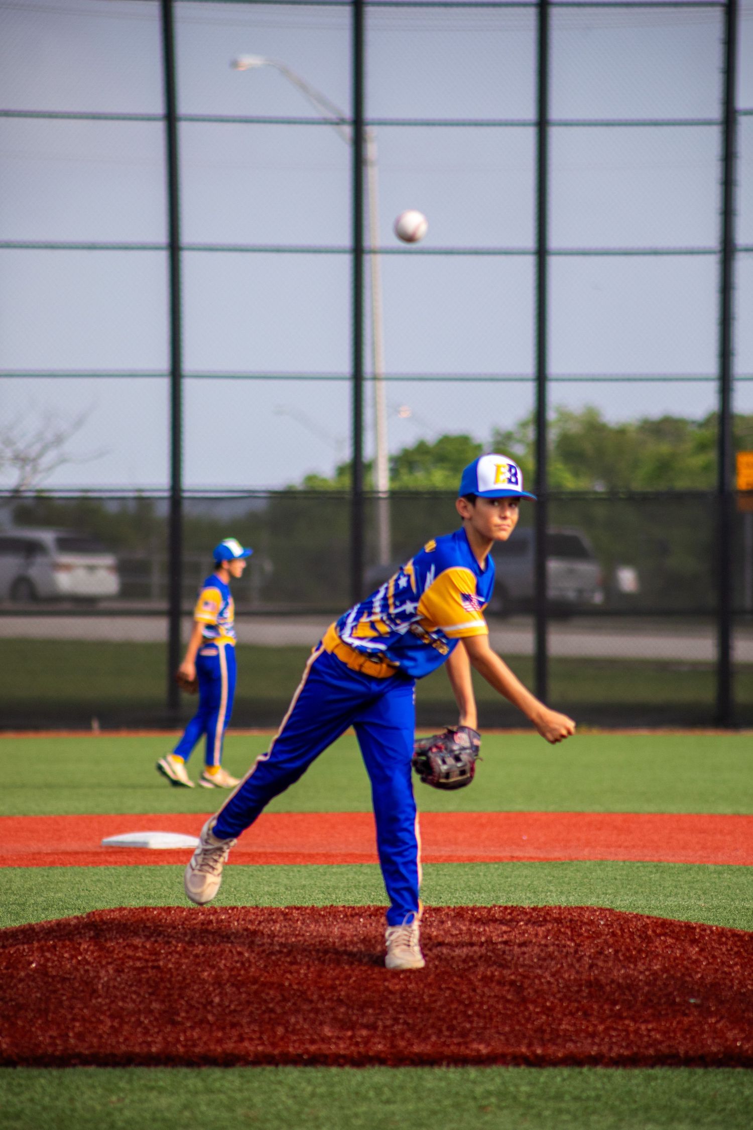 Youth baseball pitcher in blue and gold uniform mid-throw on red turf mound during a little league game.