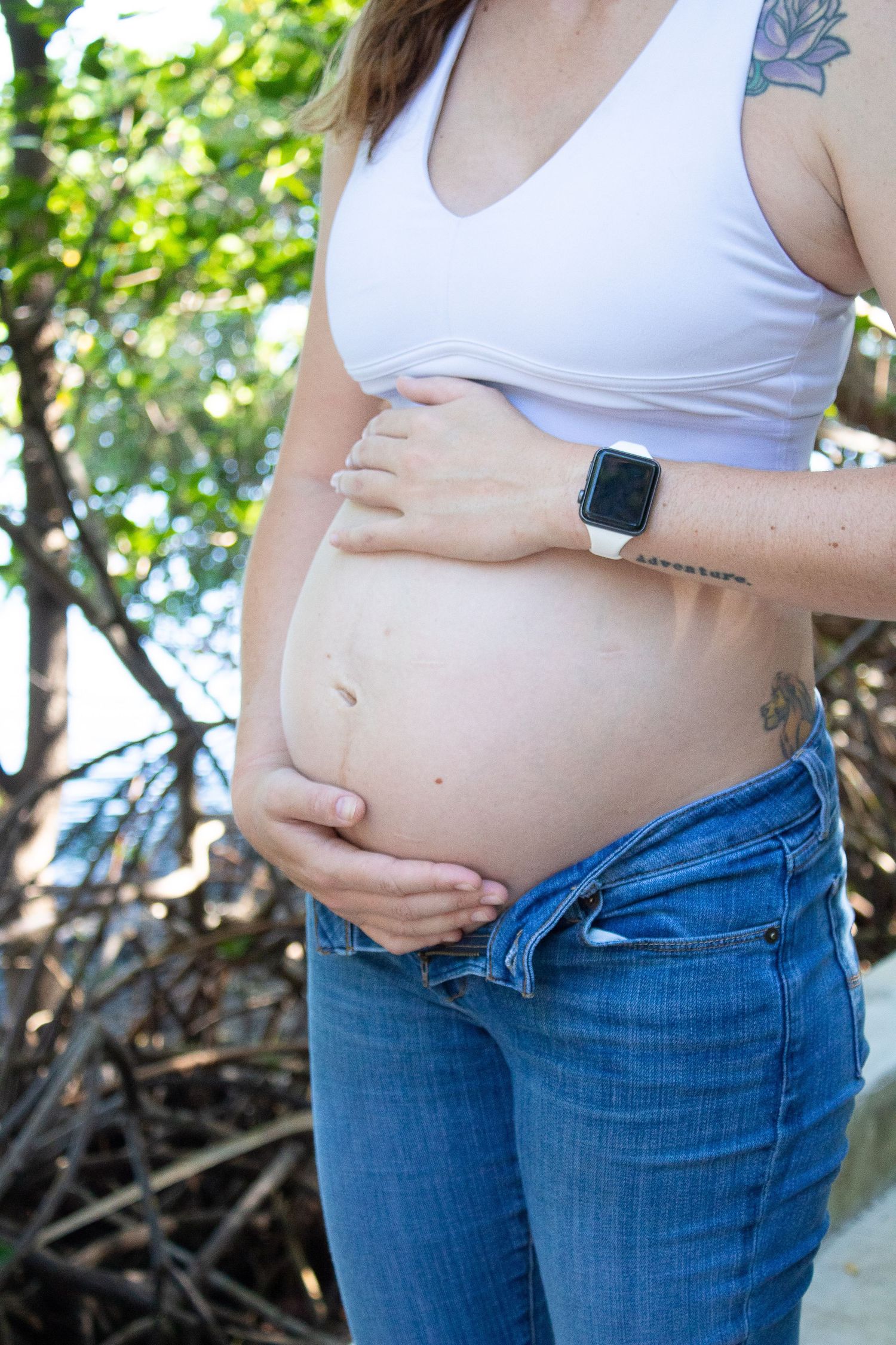 Pregnant woman in white top and jeans showing belly while wearing smartwatch.