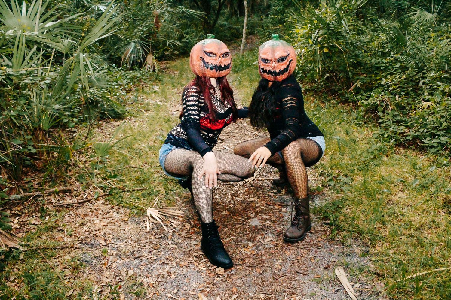 Two figures wearing pumpkin masks sit together on a log in a green forest setting.