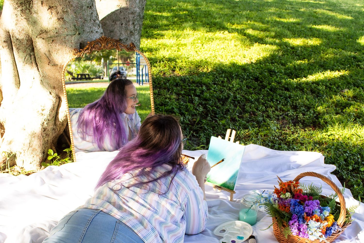 Outdoor art setup with painting supplies and a colorful wicker basket on a white blanket.