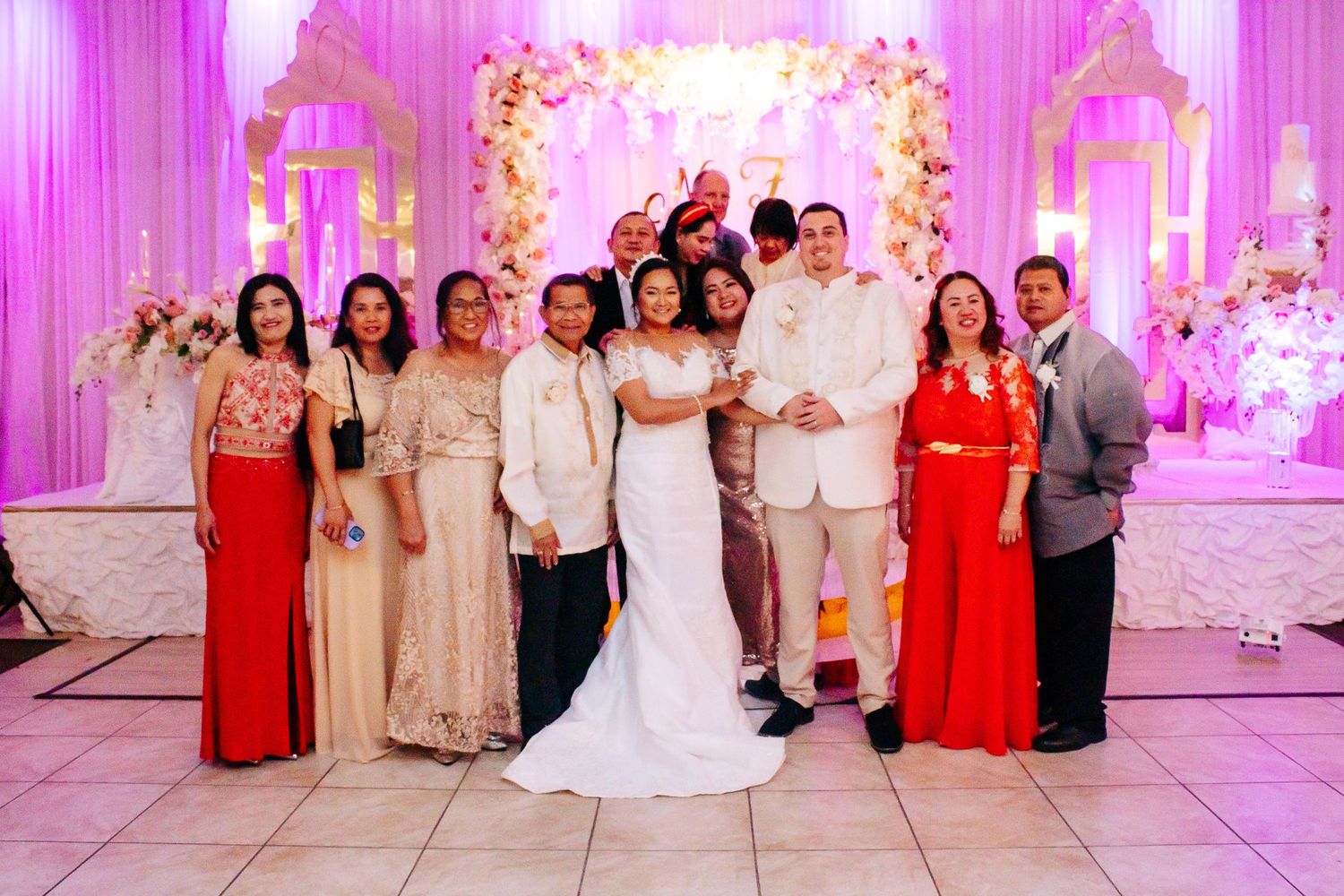 Wedding party group photo with bridesmaids in red and cream dresses against pink floral backdrop.