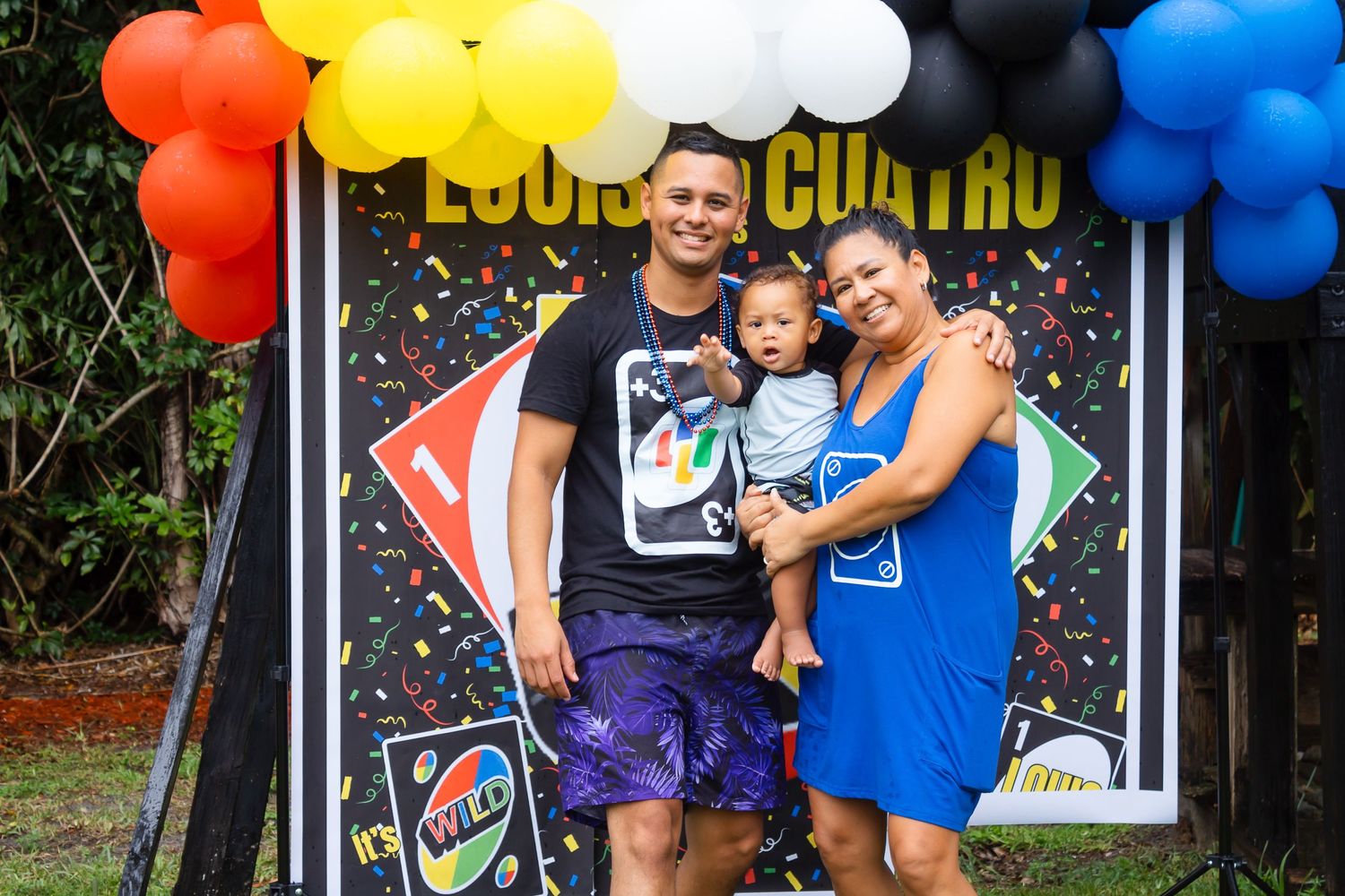 Family poses in front of colorful balloon arch and confetti backdrop at outdoor celebration.