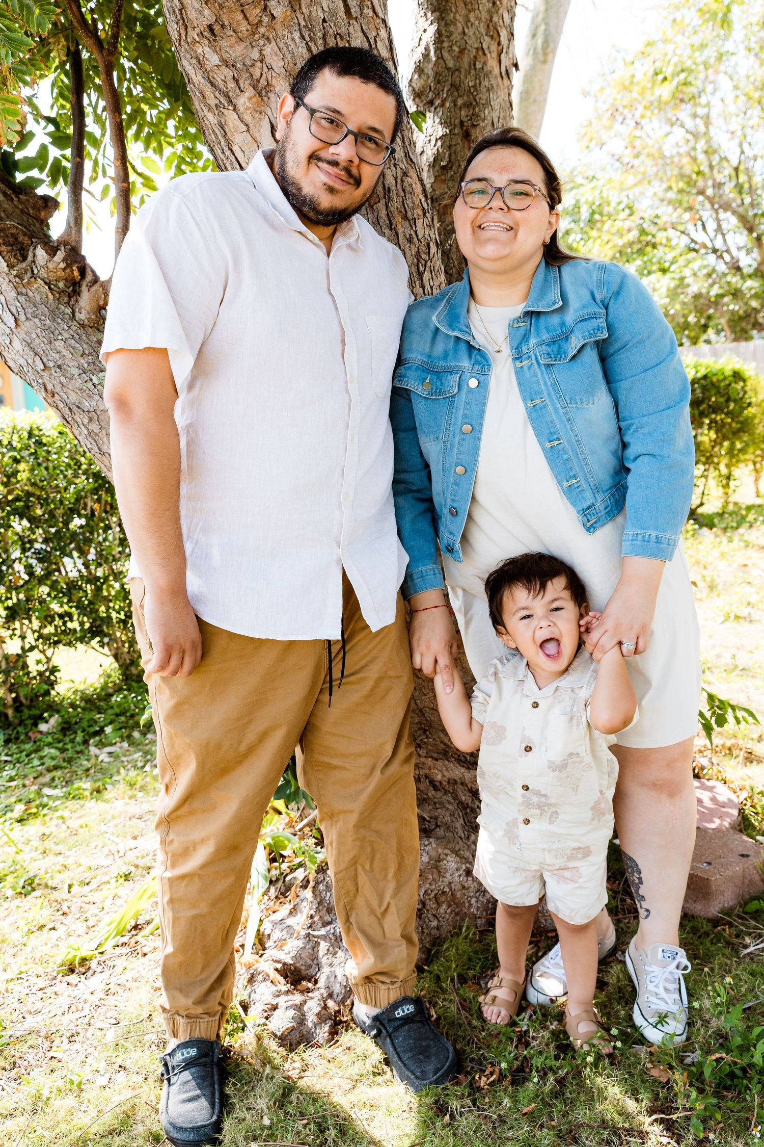 A family poses together by a tree trunk on a sunny day in casual attire.
