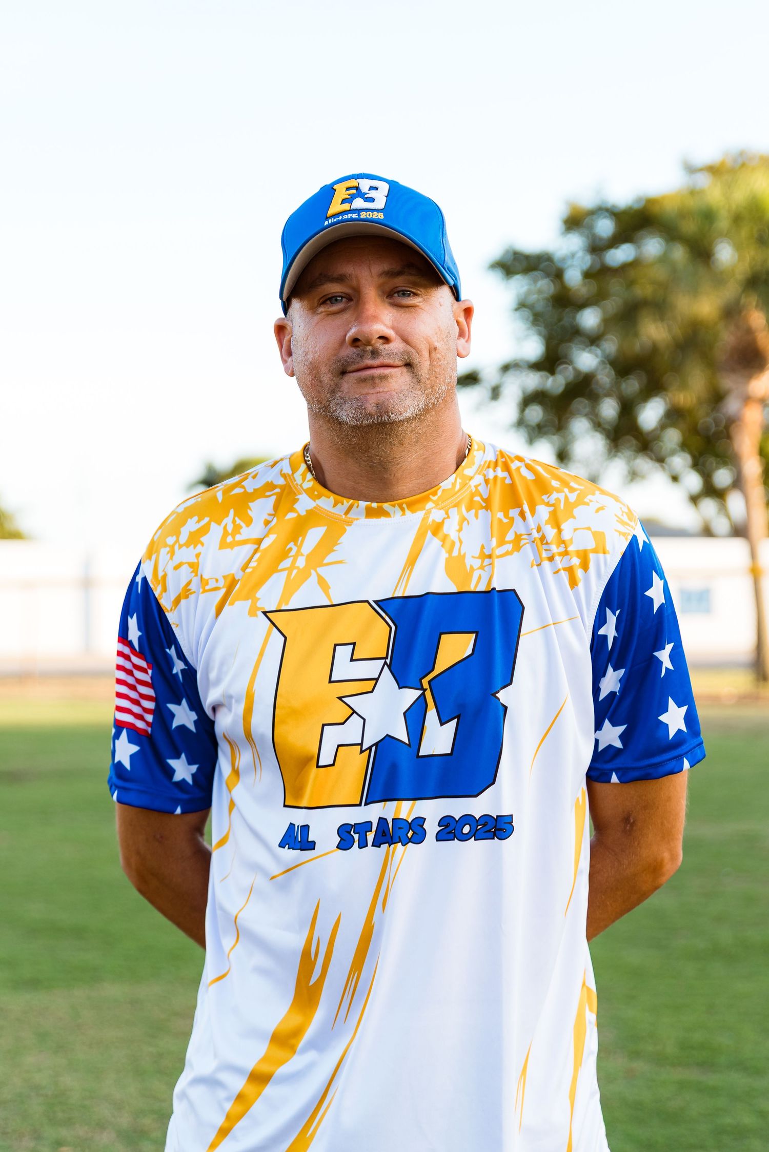 Baseball player wearing white and blue jersey with stars and stripes design posing outdoors on field.