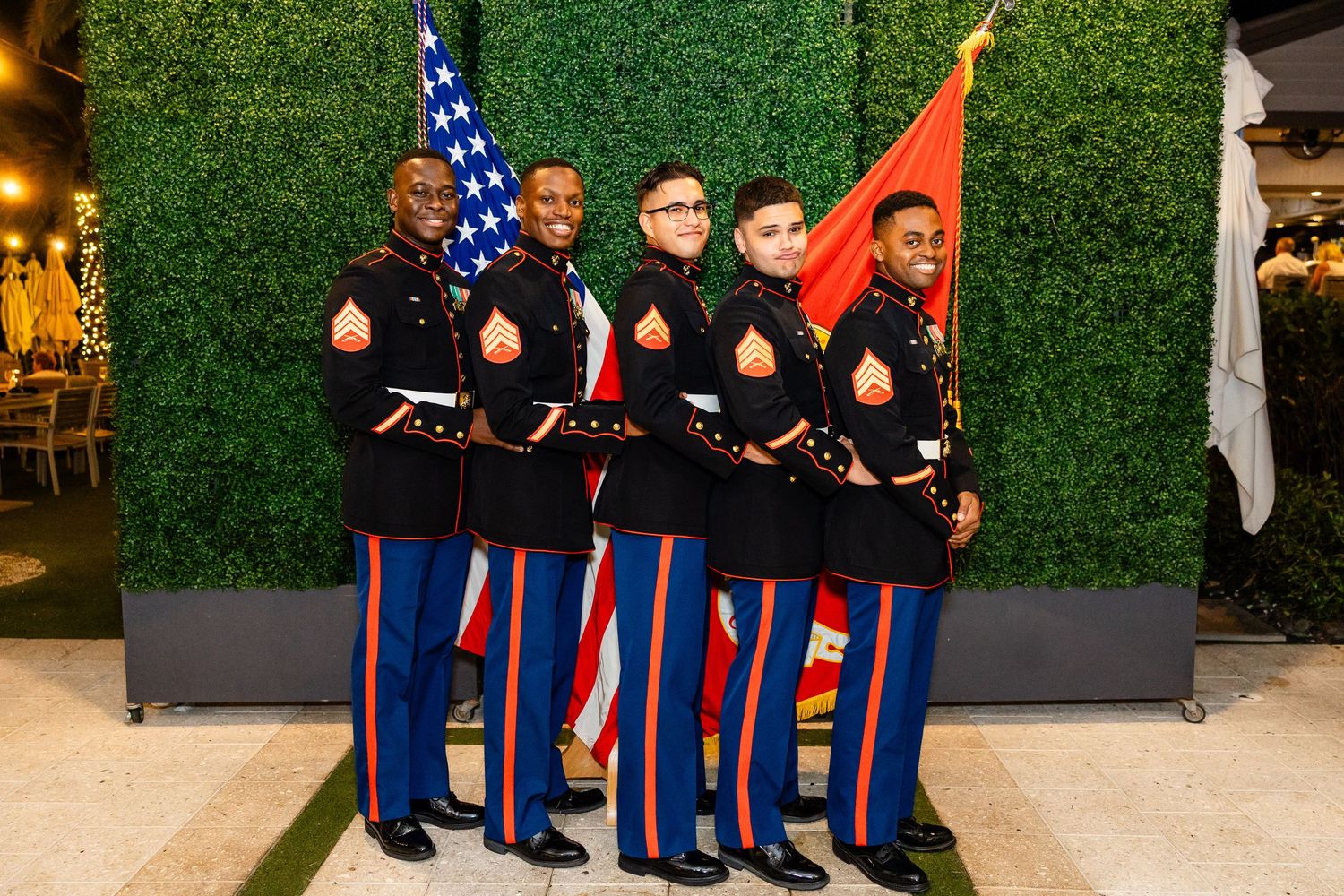 Military service members in dress uniforms stand together with American and Marine Corps flags against a green backdrop.