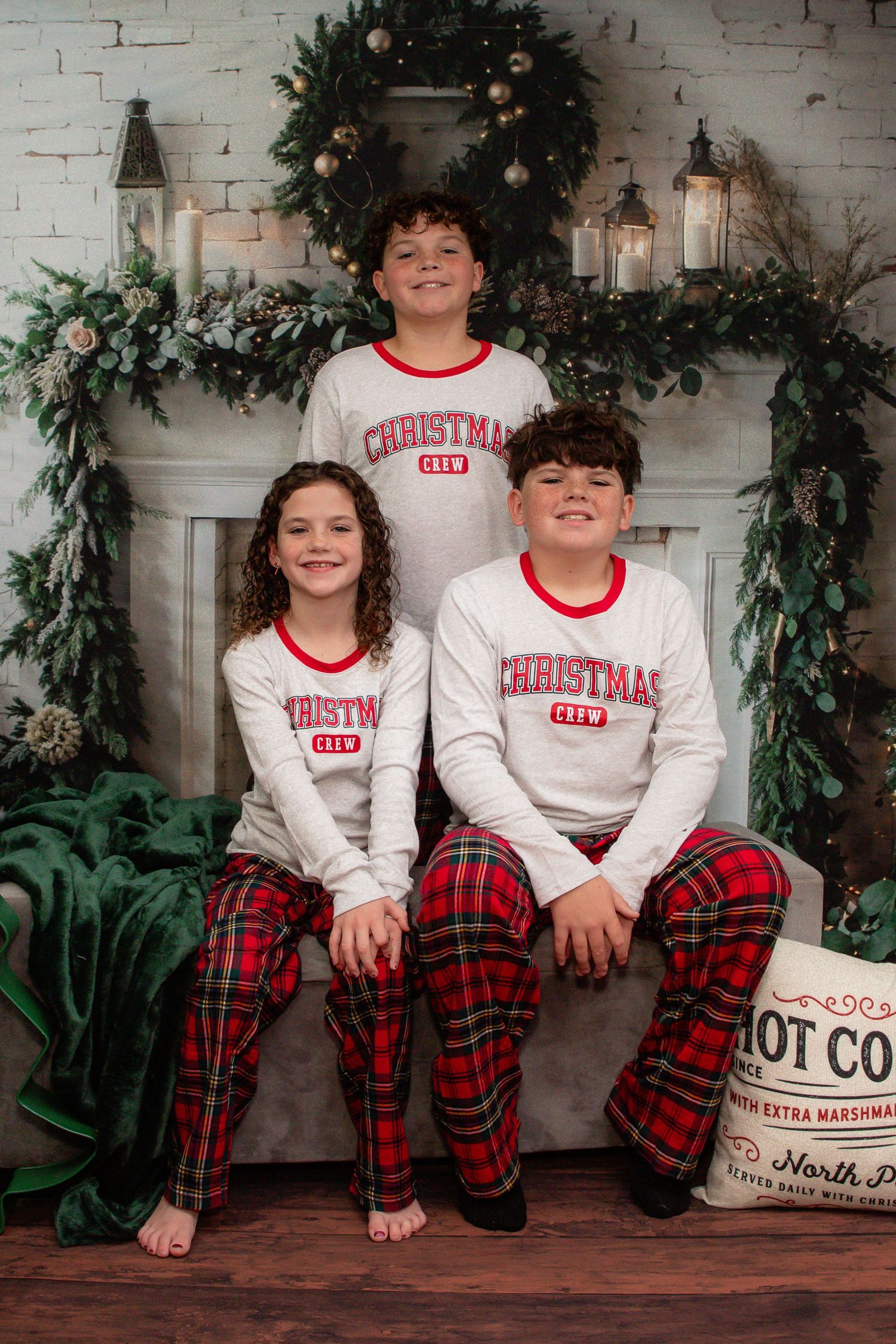 Children wearing matching Christmas pajamas with plaid pants pose against white brick wall with holiday greenery decor.