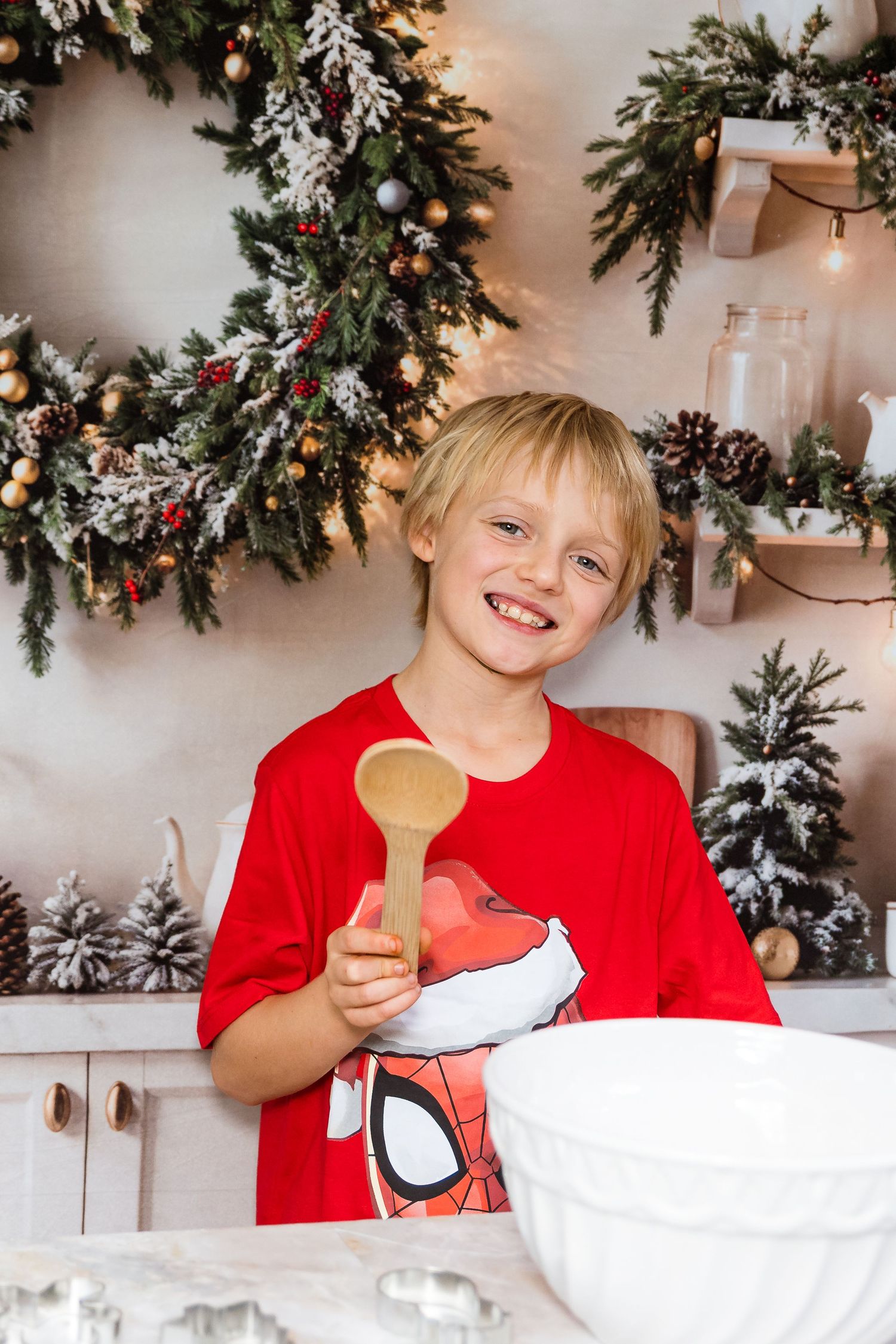 A child in a red shirt enjoys baking cookies in a festive kitchen decorated with holiday garlands and greenery.