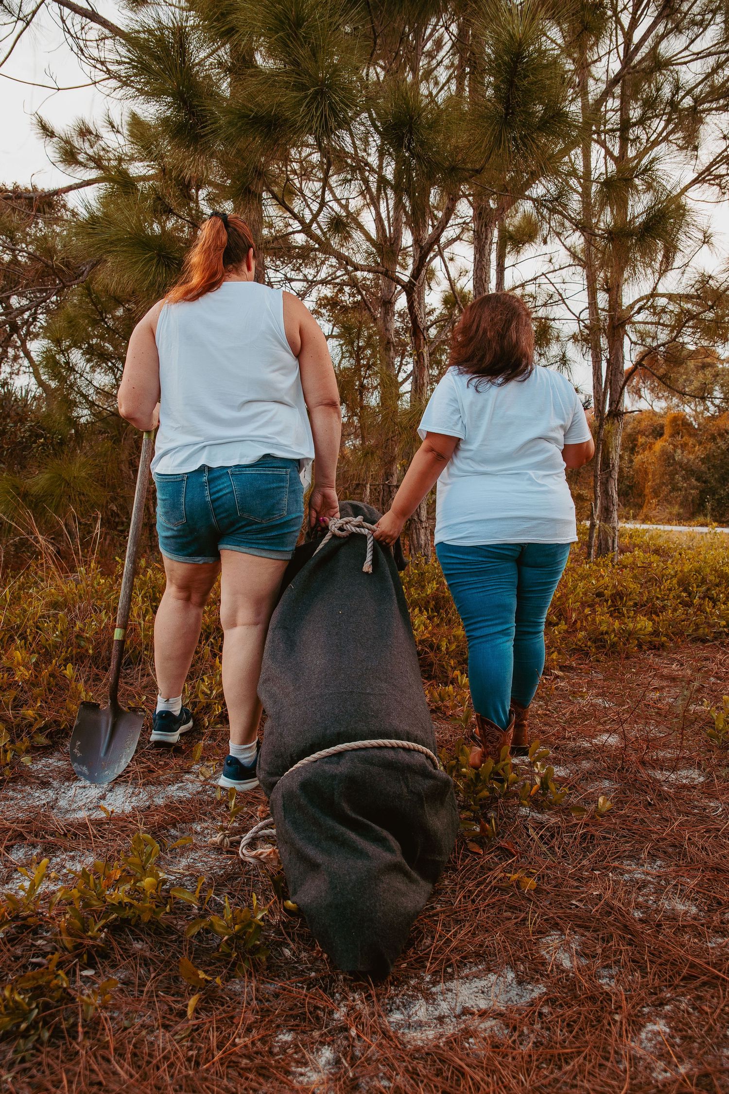 Two people in light colored shirts carry tools and equipment while walking on a nature trail among pine trees.