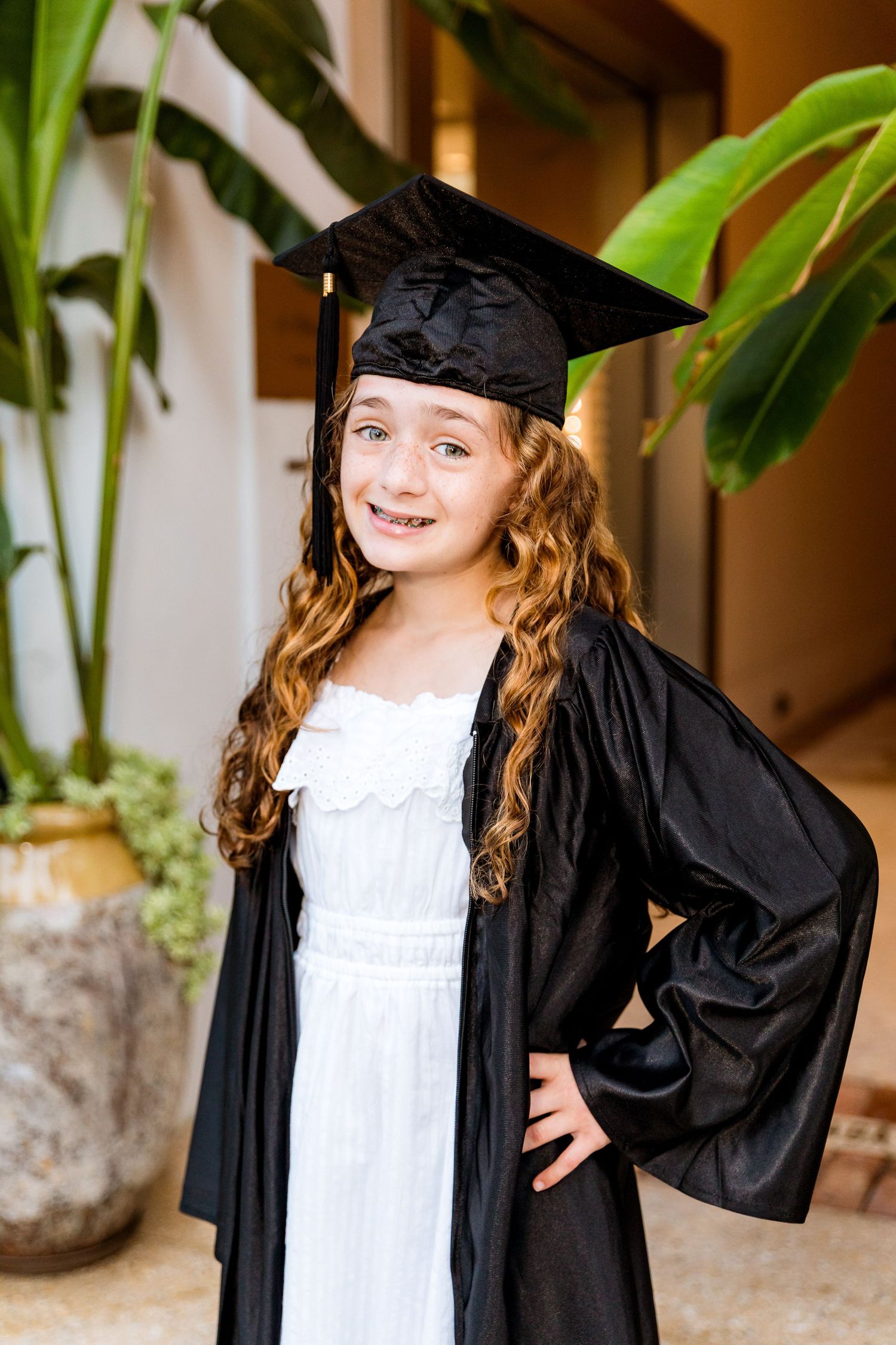 Young girl in black graduation cap and gown smiling proudly, wearing white dress with curly hair.